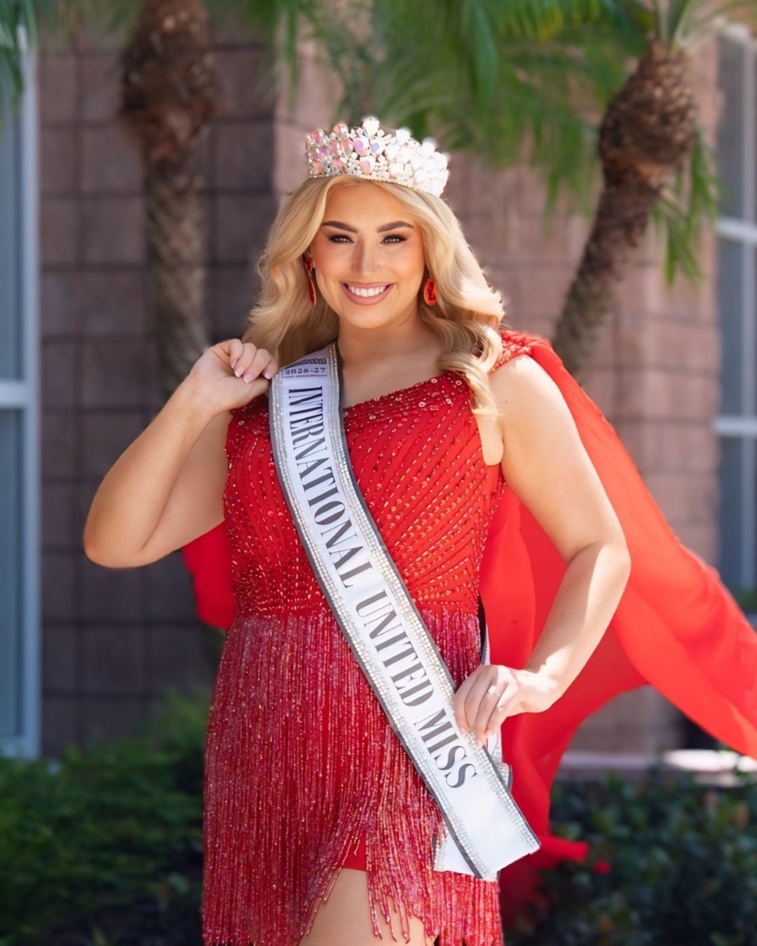 Miss International Woman wearing an orange gown, a crown, and a white sash that reads 'Miss International Woman 2025-26'. She is smiling and posing with hands on hips.