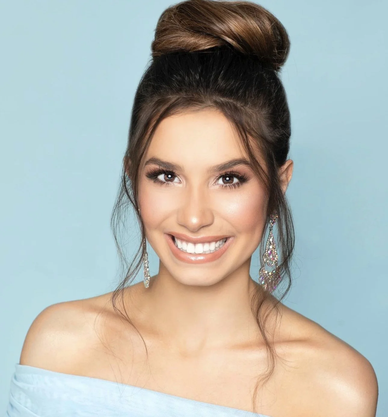 A young woman with dark brown hair styled in an elegant updo, smiling with white teeth, wearing colorful earrings, and a light blue off-the-shoulder top against a light blue background.