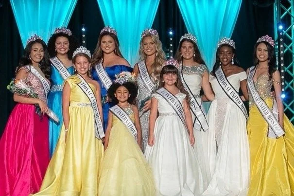 Group of nine young women and girls in crowns and sashes, dressed in colorful evening gowns, standing on a stage with blue curtains and stage lighting.