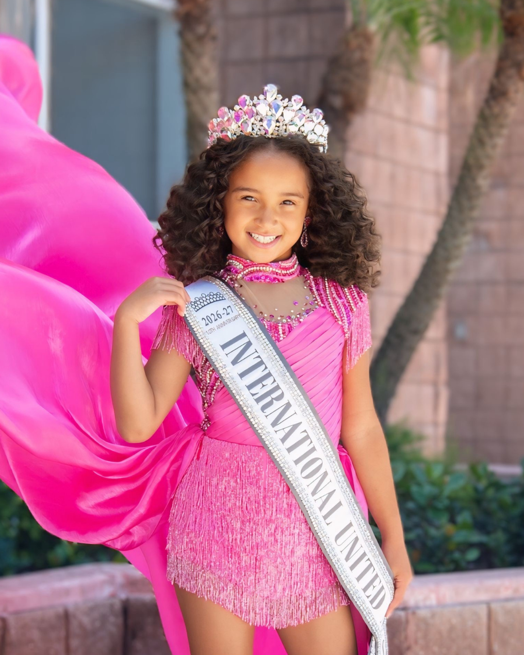 A young girl wearing a pink dress, a crown, and a sash that reads 'International United' standing with her hands on her hips.