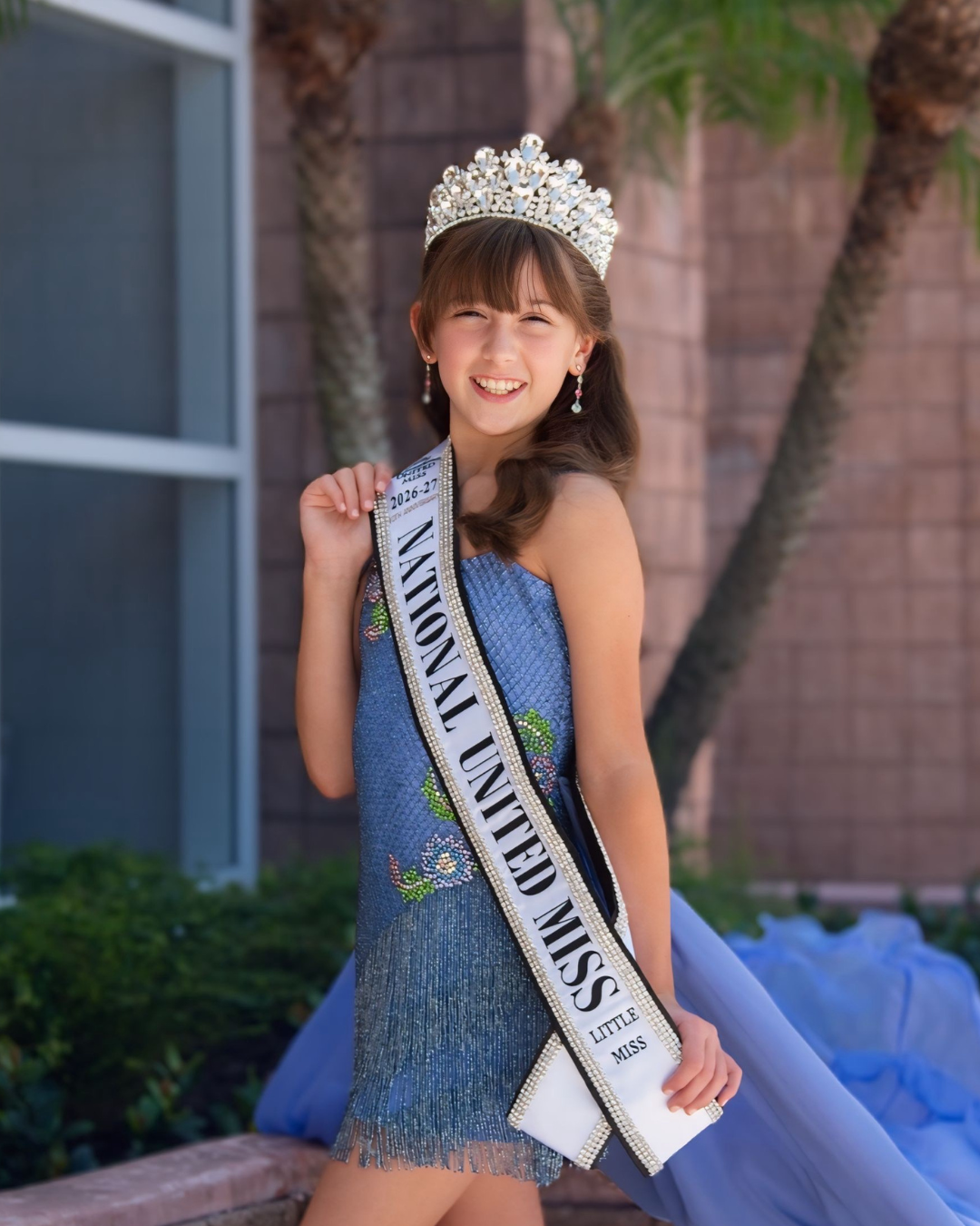 Young girl smiling wearing a blue dress, a crown, and a sash that reads 'National United Miss 2025-26'.