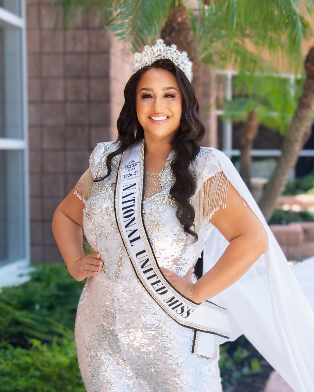 Woman wearing a crown and sash that reads Miss National United Miss 2025-26, dressed in a red gown with intricate beadwork.