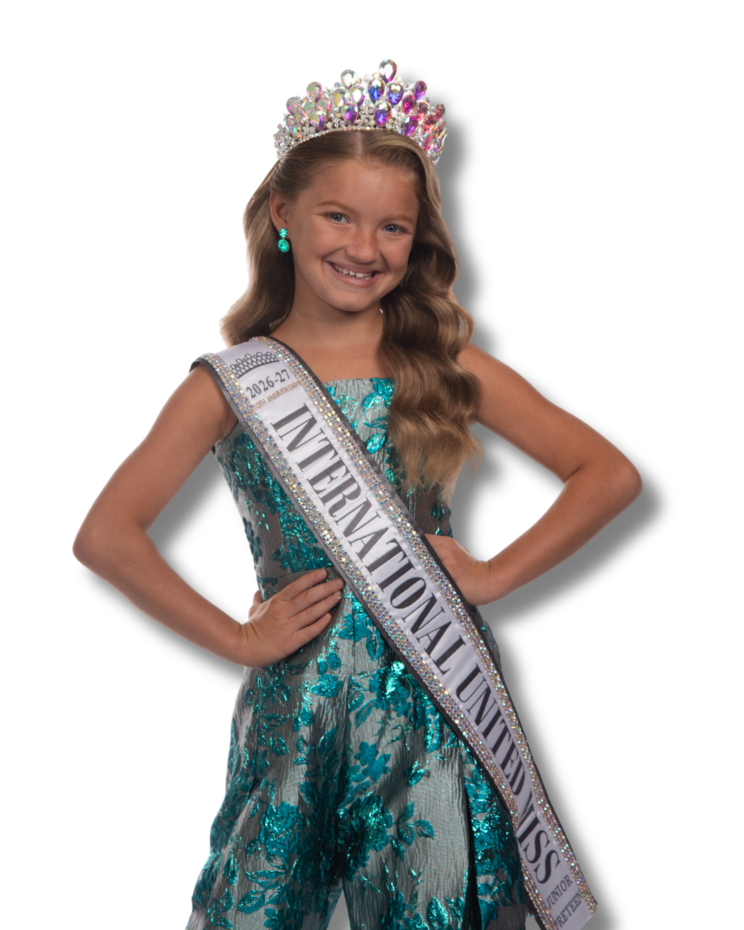 Young girl with a crown, earrings, and a sash that reads "International United Miss" wearing a glittering turquoise gown with a tulle skirt and pose with her hands on her hips, smiling.