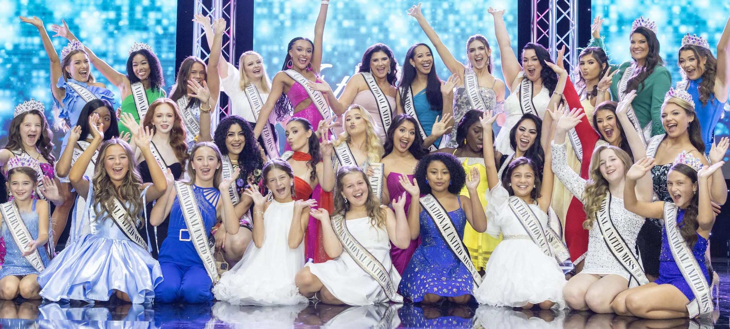 Group of young women and girls on stage celebrating winners of a beauty pageant, wearing sashes and crowns, smiling and cheering.