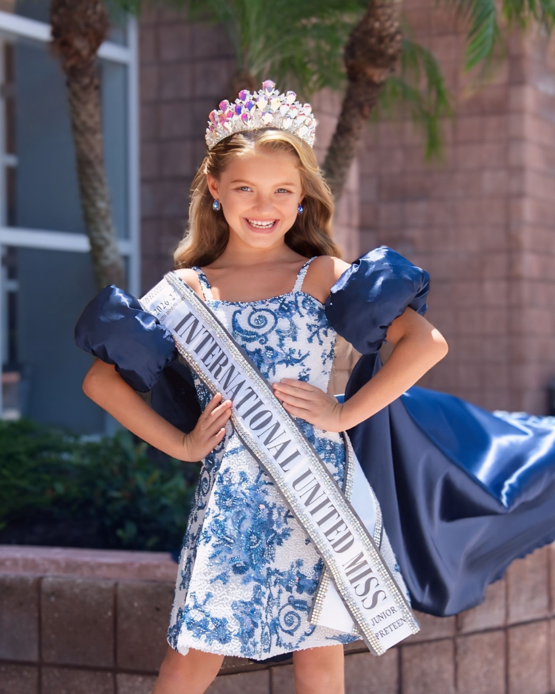 Young girl with a crown, earrings, and a sash that reads "International United Miss" wearing a glittering turquoise gown with a tulle skirt and pose with her hands on her hips, smiling.