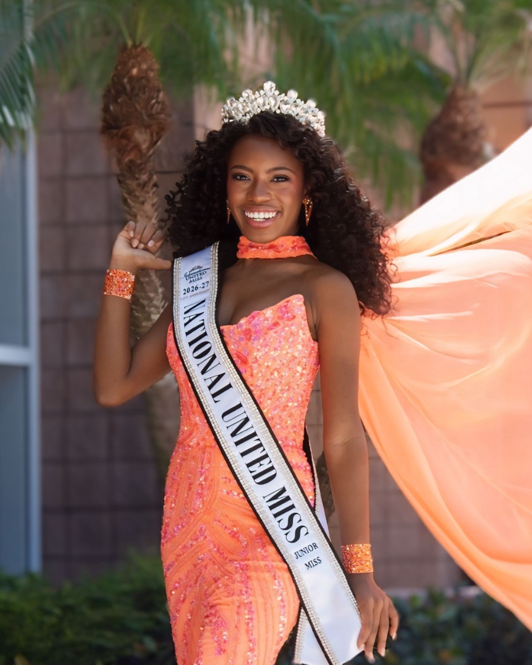 Young woman wearing a blue evening gown, a sash labeled 'National United Miss 2025-26,' and a sparkling tiara, smiling with hands on her hips.