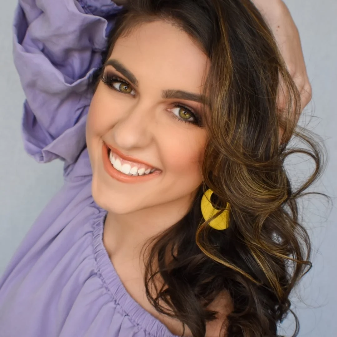 A woman with wavy brown hair and green eyes smiling, wearing a lavender top and yellow earrings.