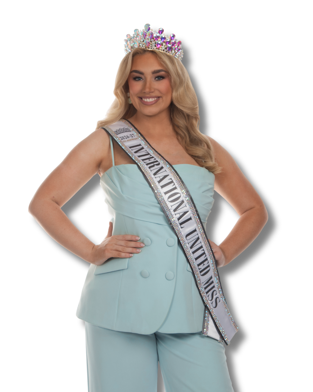 Miss International Woman wearing an orange gown, a crown, and a white sash that reads 'Miss International Woman 2025-26'. She is smiling and posing with hands on hips.