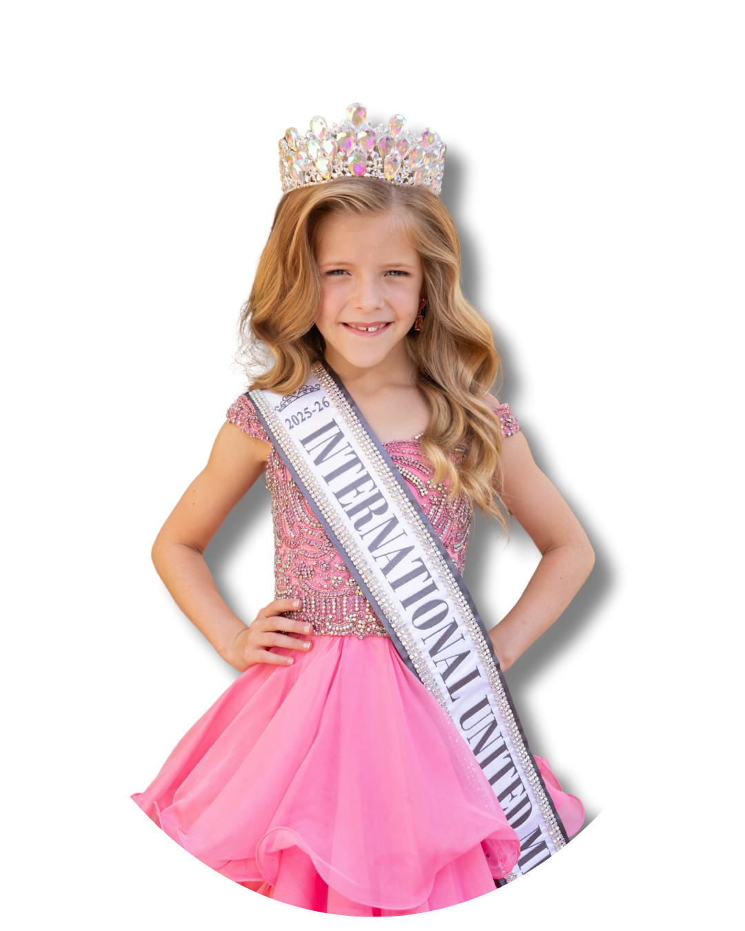 A young girl wearing a pink dress, a crown, and a sash that reads 'International United' standing with her hands on her hips.