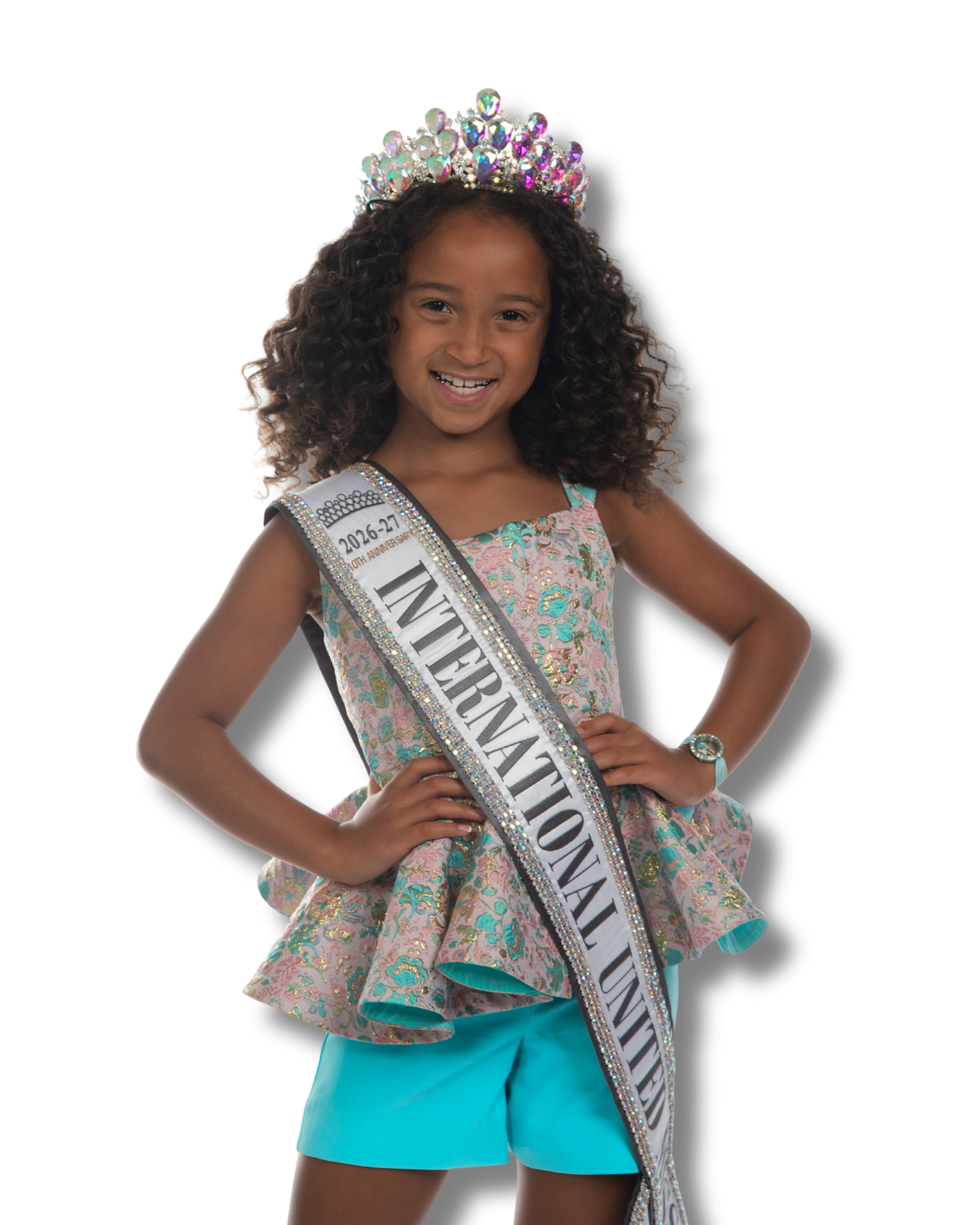 A young girl wearing a pink dress, a crown, and a sash that reads 'International United' standing with her hands on her hips.