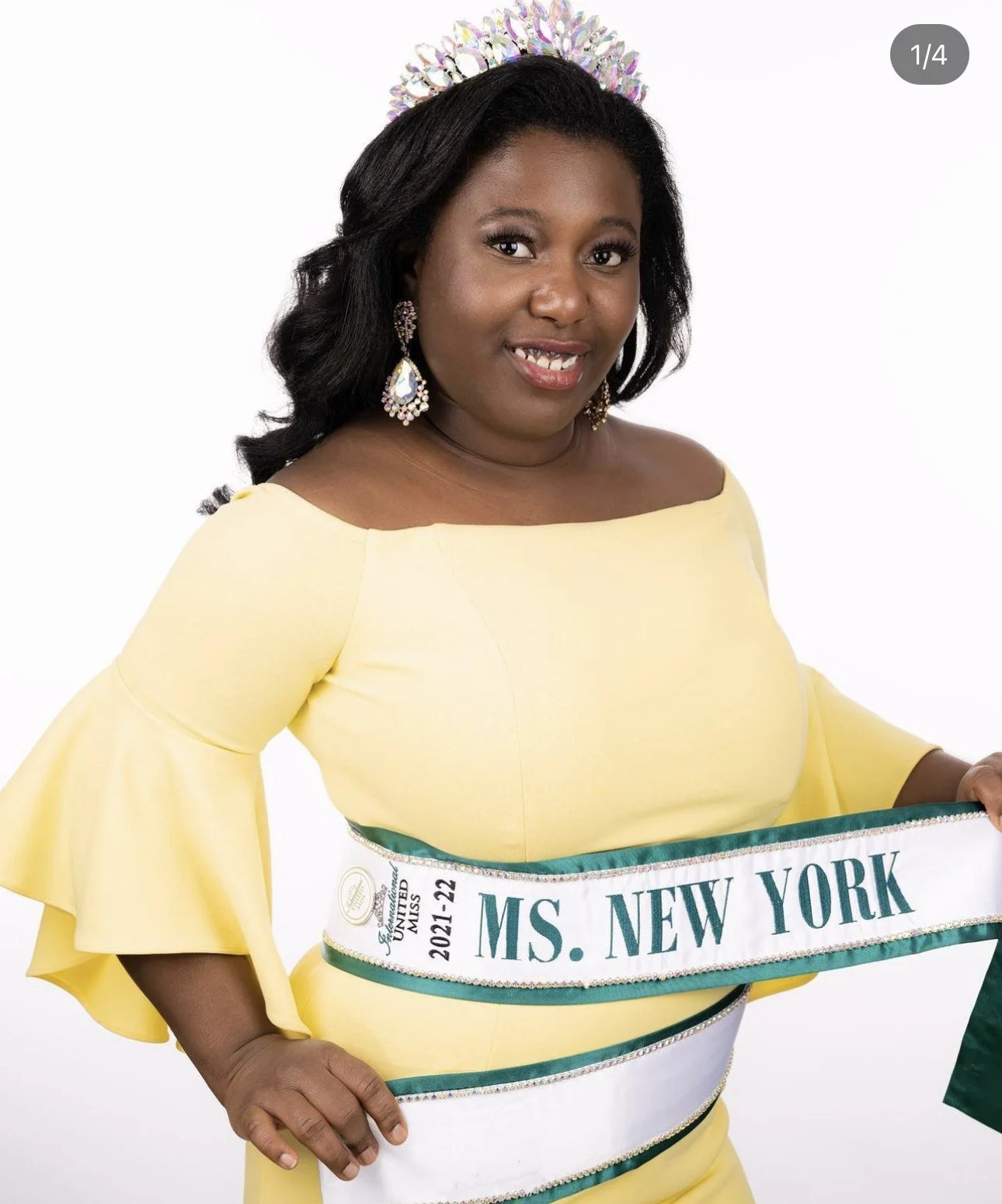 A woman wearing a yellow off-the-shoulder dress, a sparkling tiara, and large earrings, holding a sash that reads "MS. NEW YORK" and mentions her participation in the Miss United States pageant 2021-22.