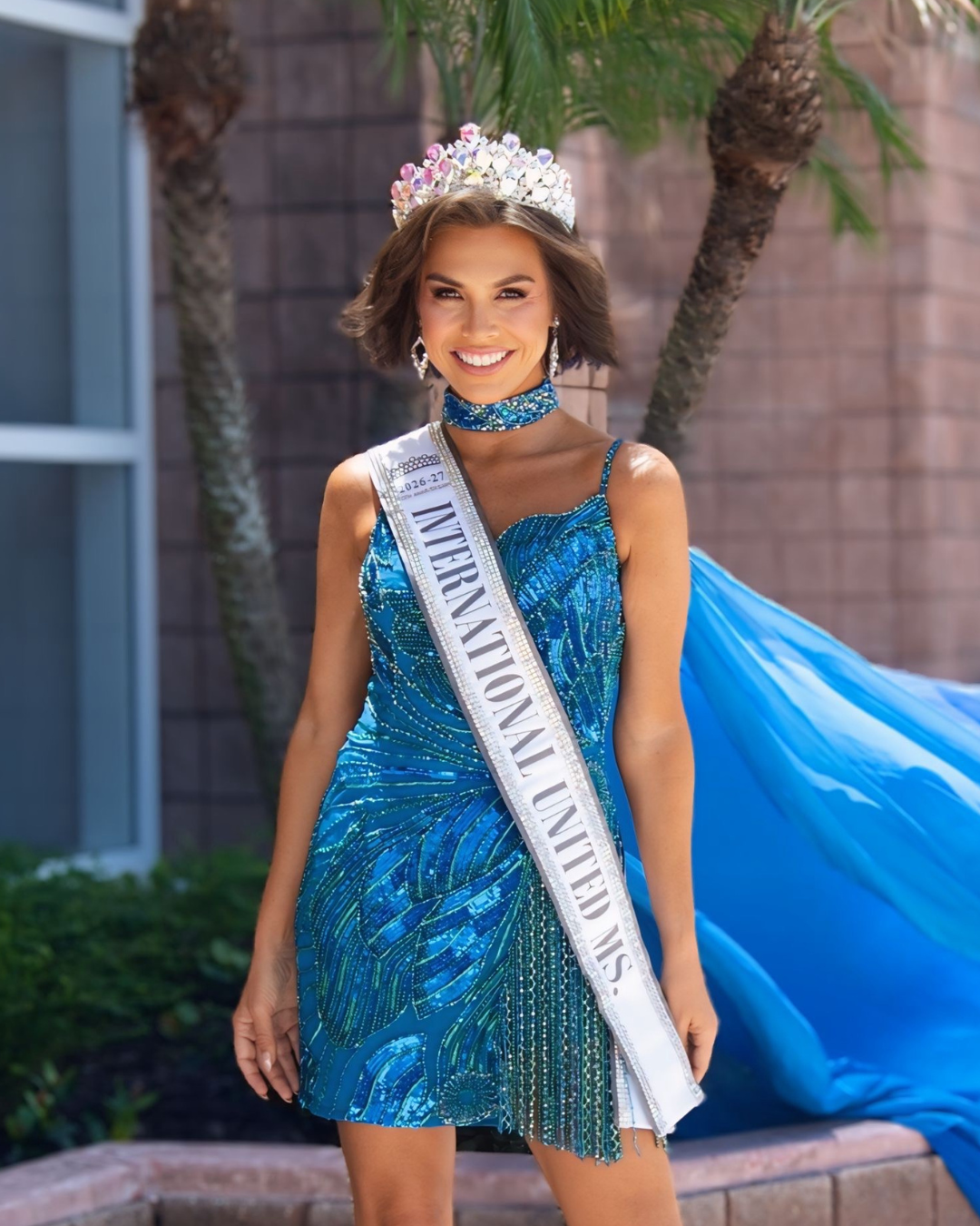 A woman wearing a purple gown, crown, and sash that reads '2025-26 INTERNATIONAL UNITED MS.' smiling with her hands on her hips.