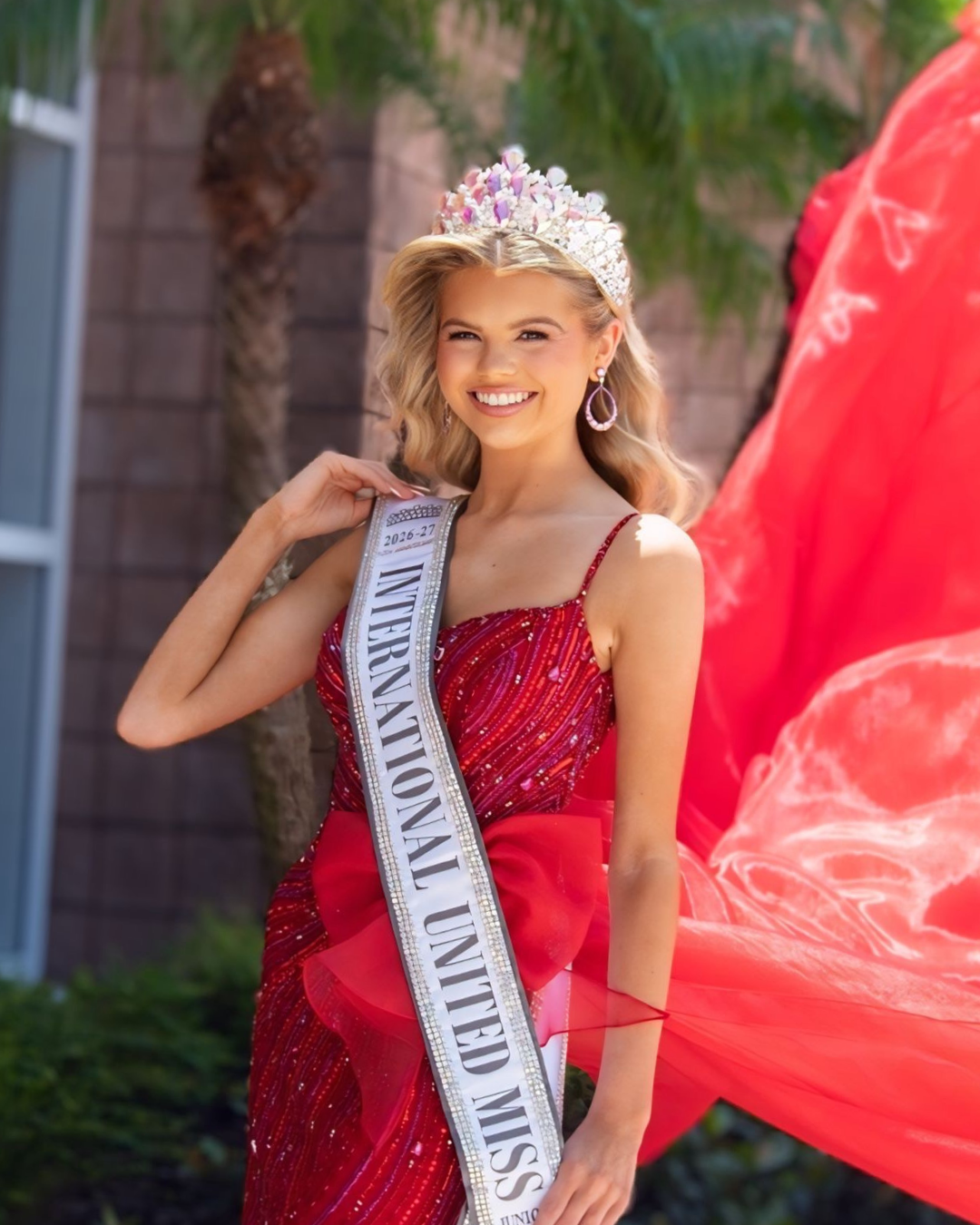 Beauty queen wearing a crown, pink gown, and sash that reads 'International United Miss 2025-26' smiling with hands on hips.