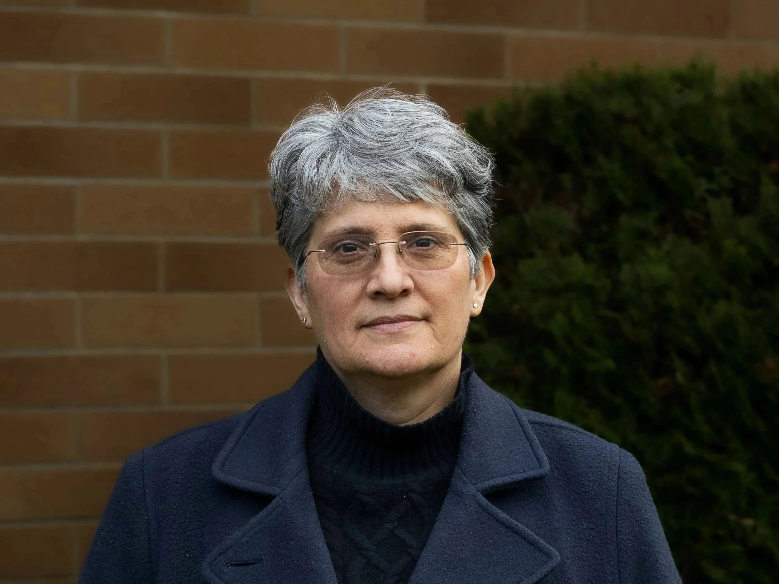 A woman in a turtleneck and jacket poses in front of a brick wall and greenery