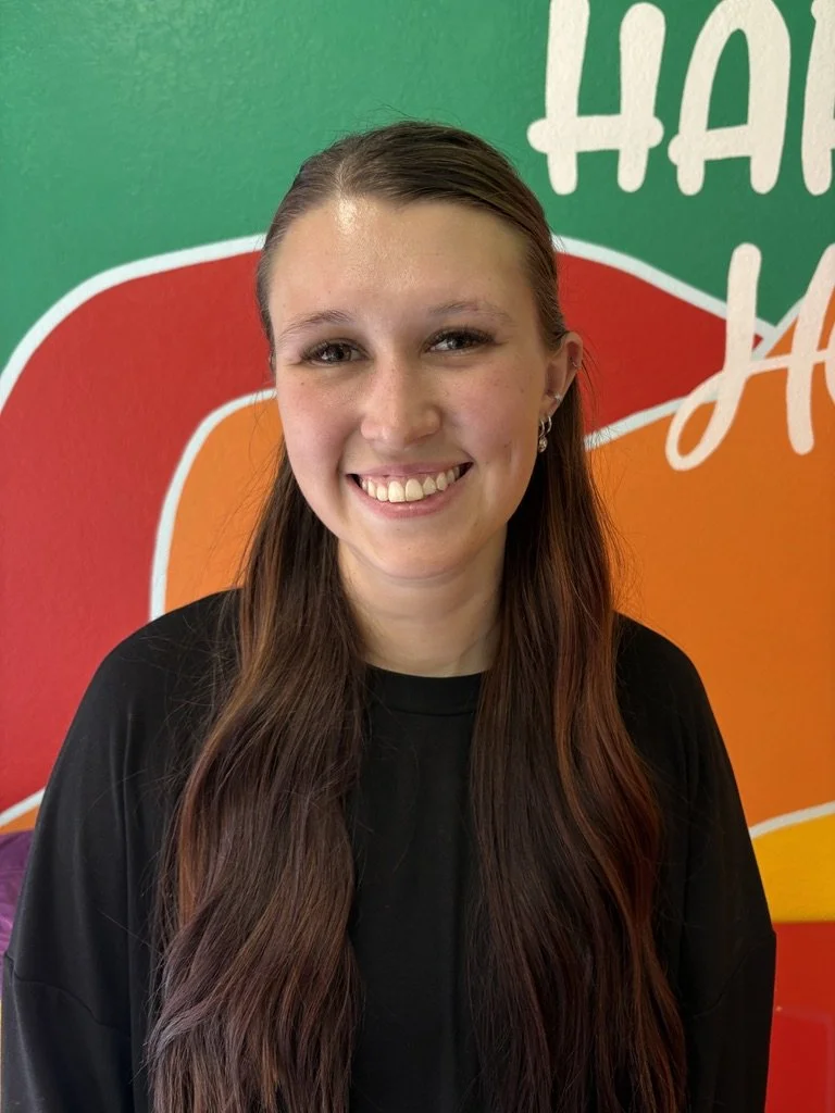 Smiling young woman with long brown hair standing in front of a colorful wall with the words "HAPPY" and "HOUR" visible.