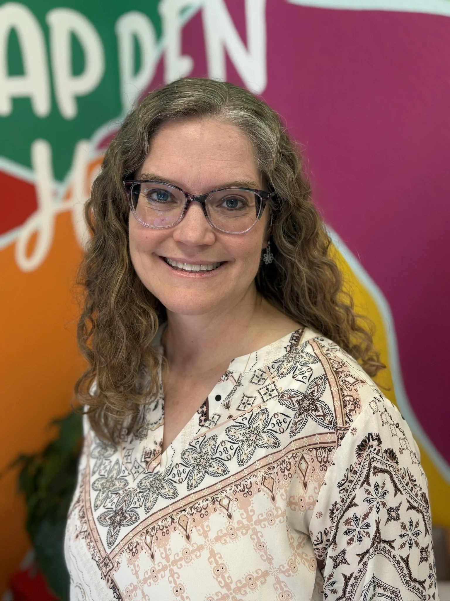 A woman smiling, wearing glasses and a white patterned shirt, standing in front of a colorful background with pink, yellow, green, and orange sections and abstract designs.