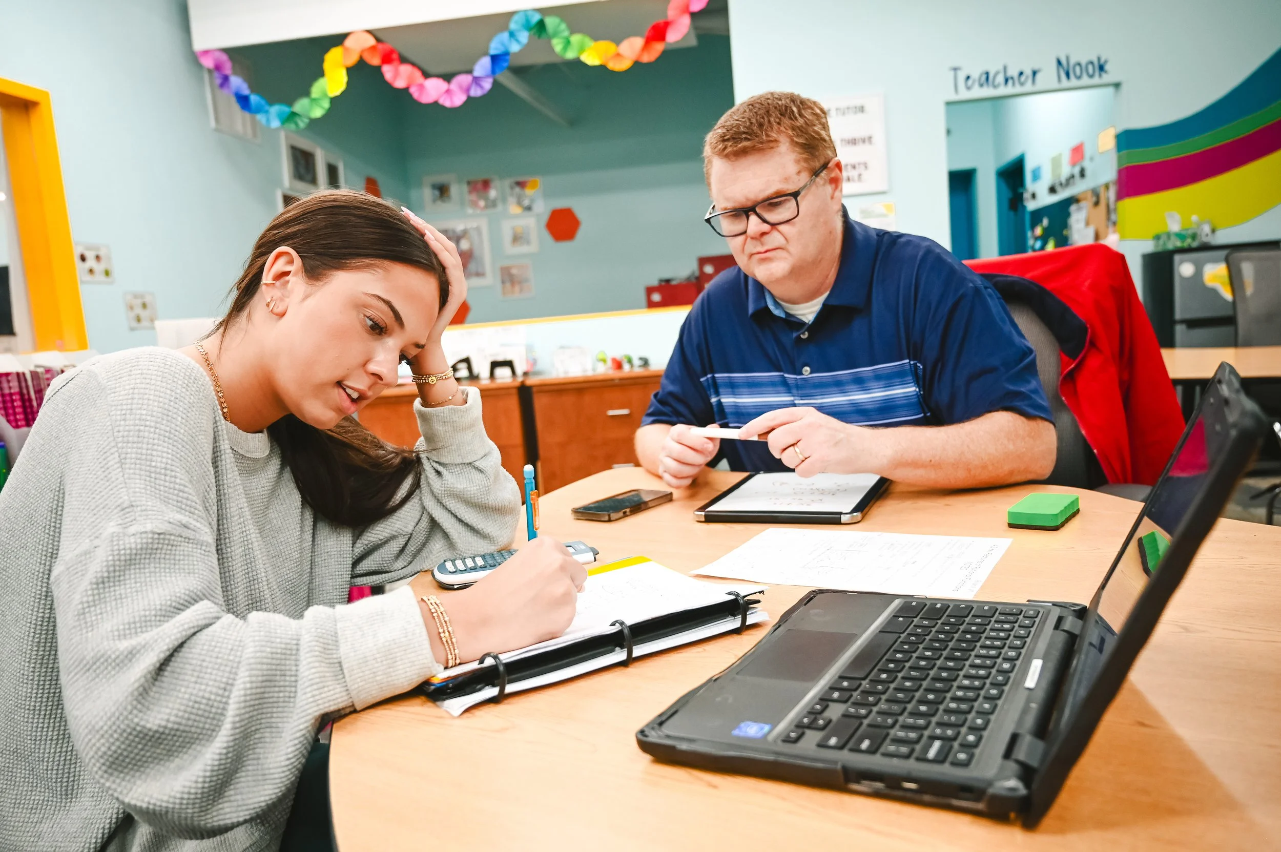 A young woman and an older man sit at a desk in a colorful classroom, working with papers, calculator, and a tablet, with decorations and signs on the walls.