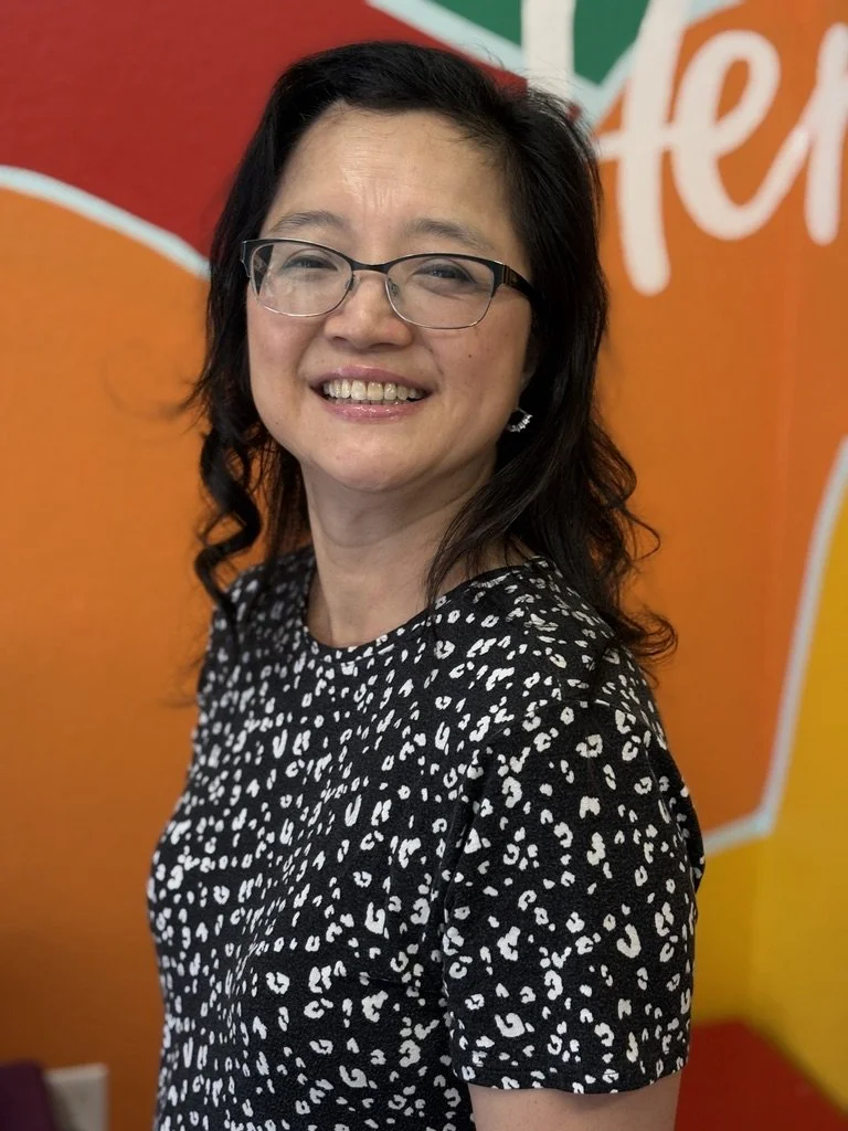 A smiling woman with black hair, glasses, and earrings, wearing a black and white patterned top, standing in front of an orange background with white and green elements.