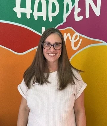 A woman with long brown hair and glasses smiling in front of a colorful mural with the words 'HAPPEN' and 'me'.