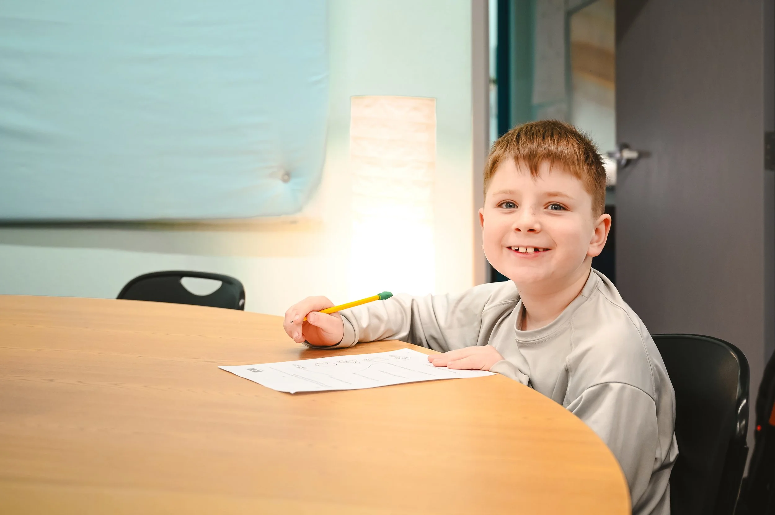 A young boy with brown hair and blue eyes smiling at the camera, sitting at a wooden table with a pencil in his right hand and a worksheet in front of him in a classroom or office setting.