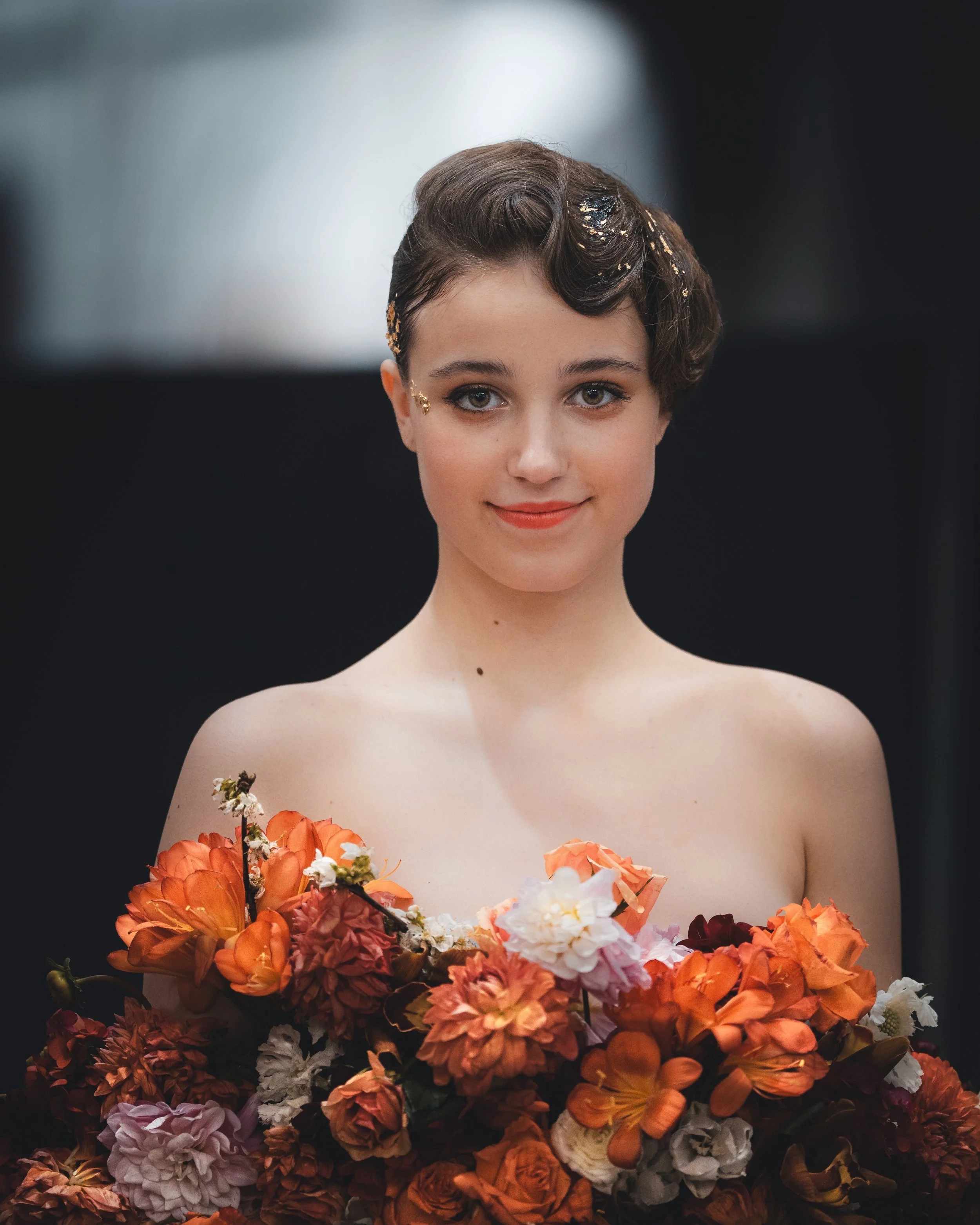 Young woman with short, styled dark hair holding a large bouquet of colorful flowers, smiling softly, with a dark background.