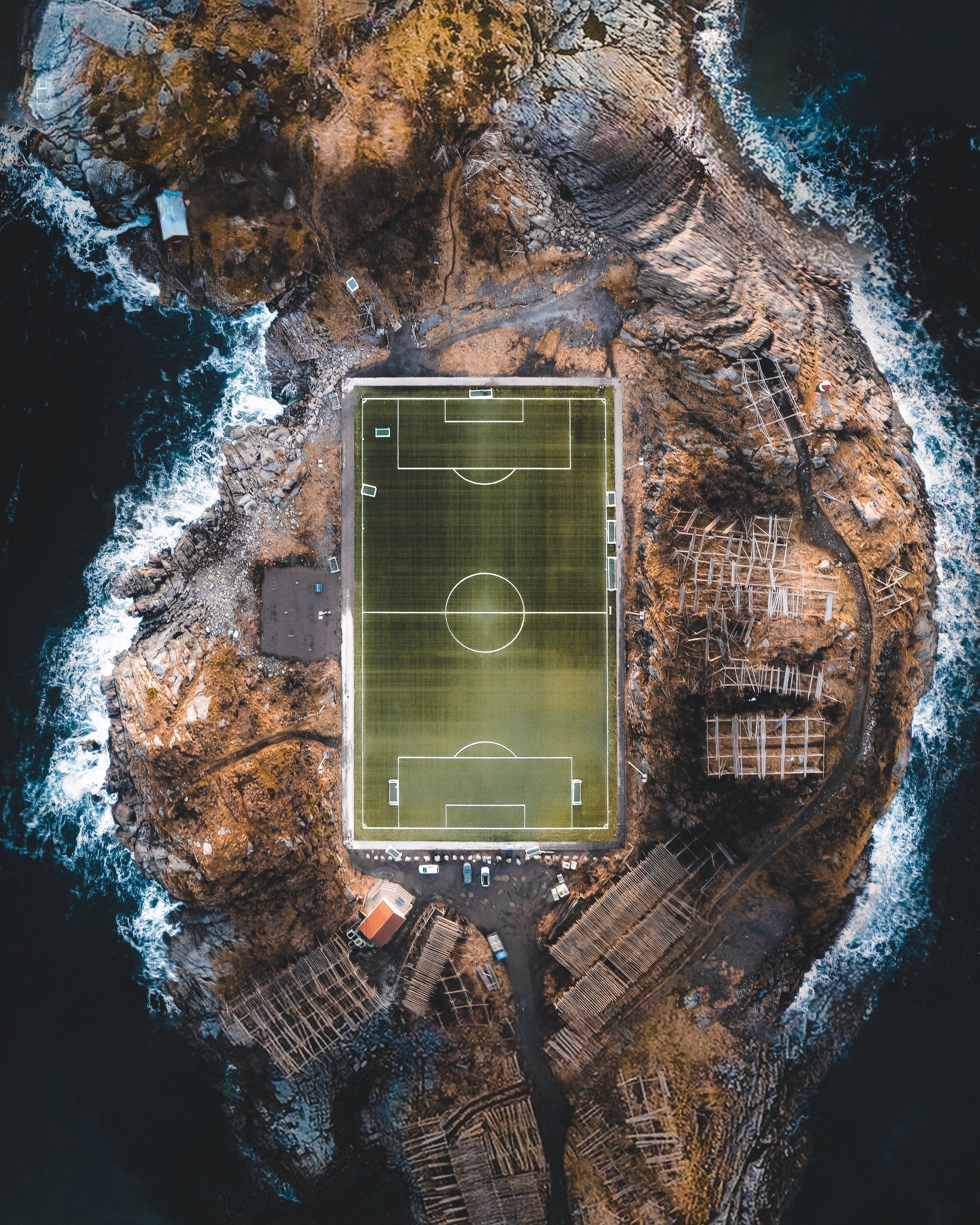 A soccer field is situated on a small rocky island surrounded by water, with construction scaffolding and pathways around it.