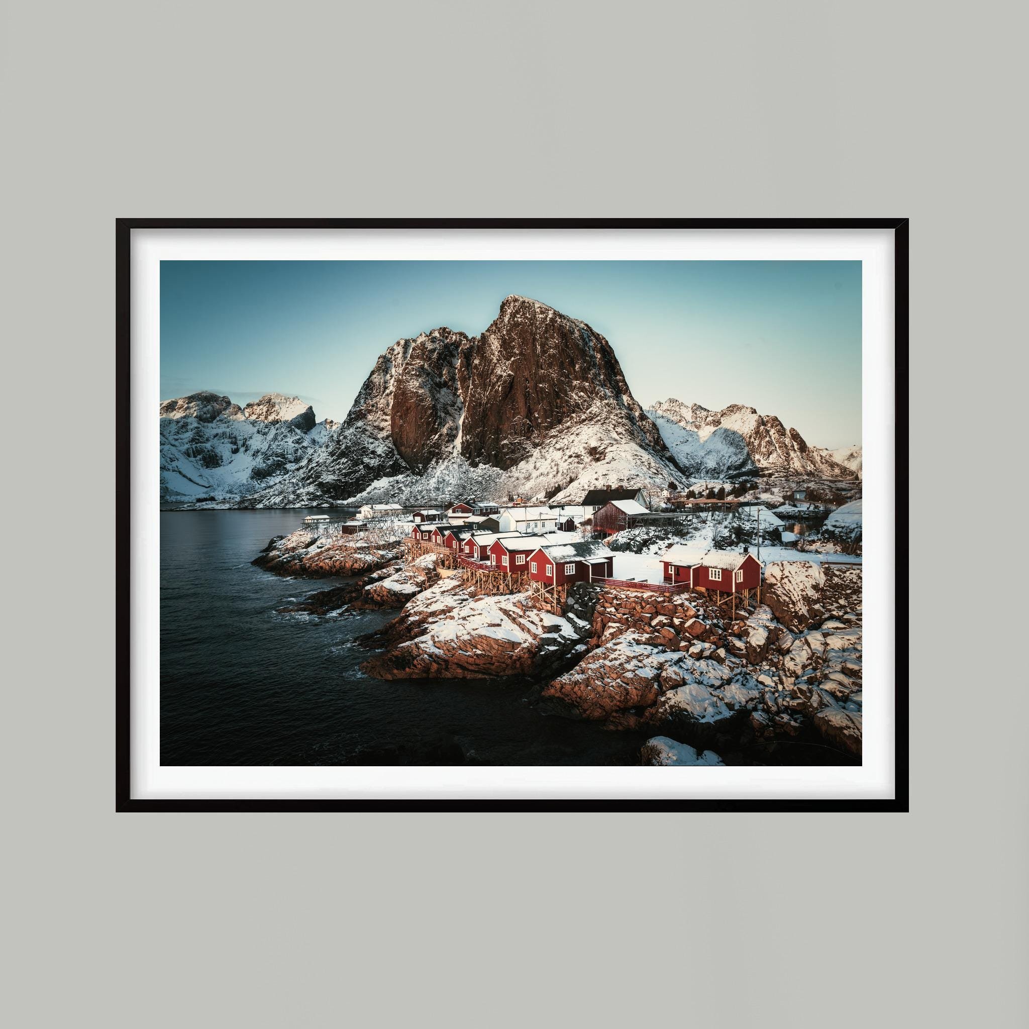 Red cabins beside dark water beneath snowy mountains in Hamnøy, Lofoten, shown in a black frame against a light grey background. Frame not included.