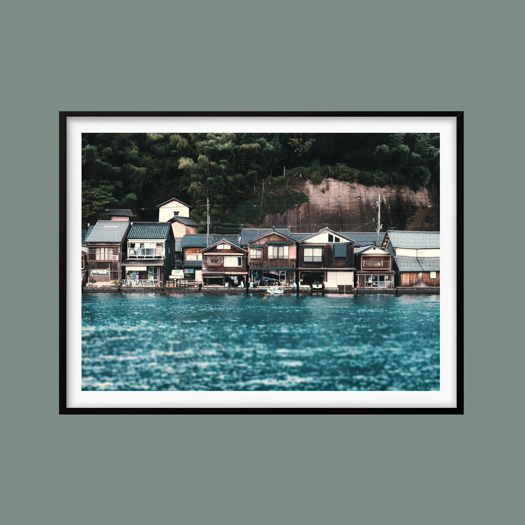 Ine Bay boathouses beside blue water, shown as a landscape photograph in a black frame against a muted green background. Frame not included.