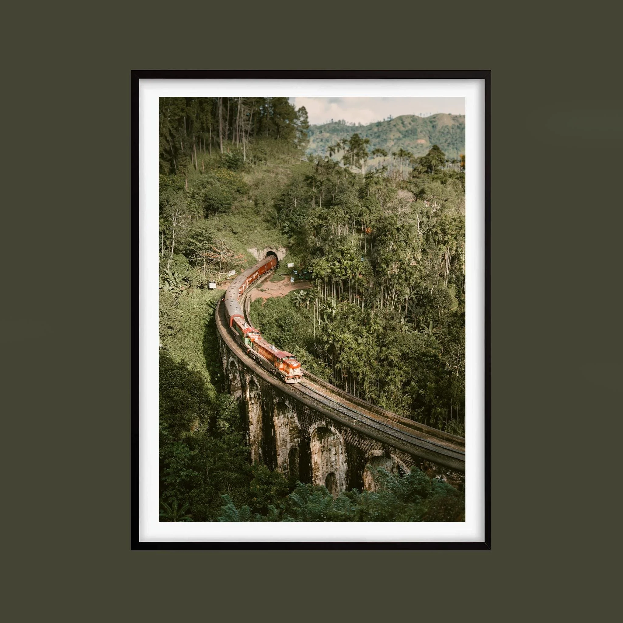 Red train crossing Nine Arches Bridge amid green hills in Ella, Sri Lanka, shown in a black frame against an olive background. Frame not included.