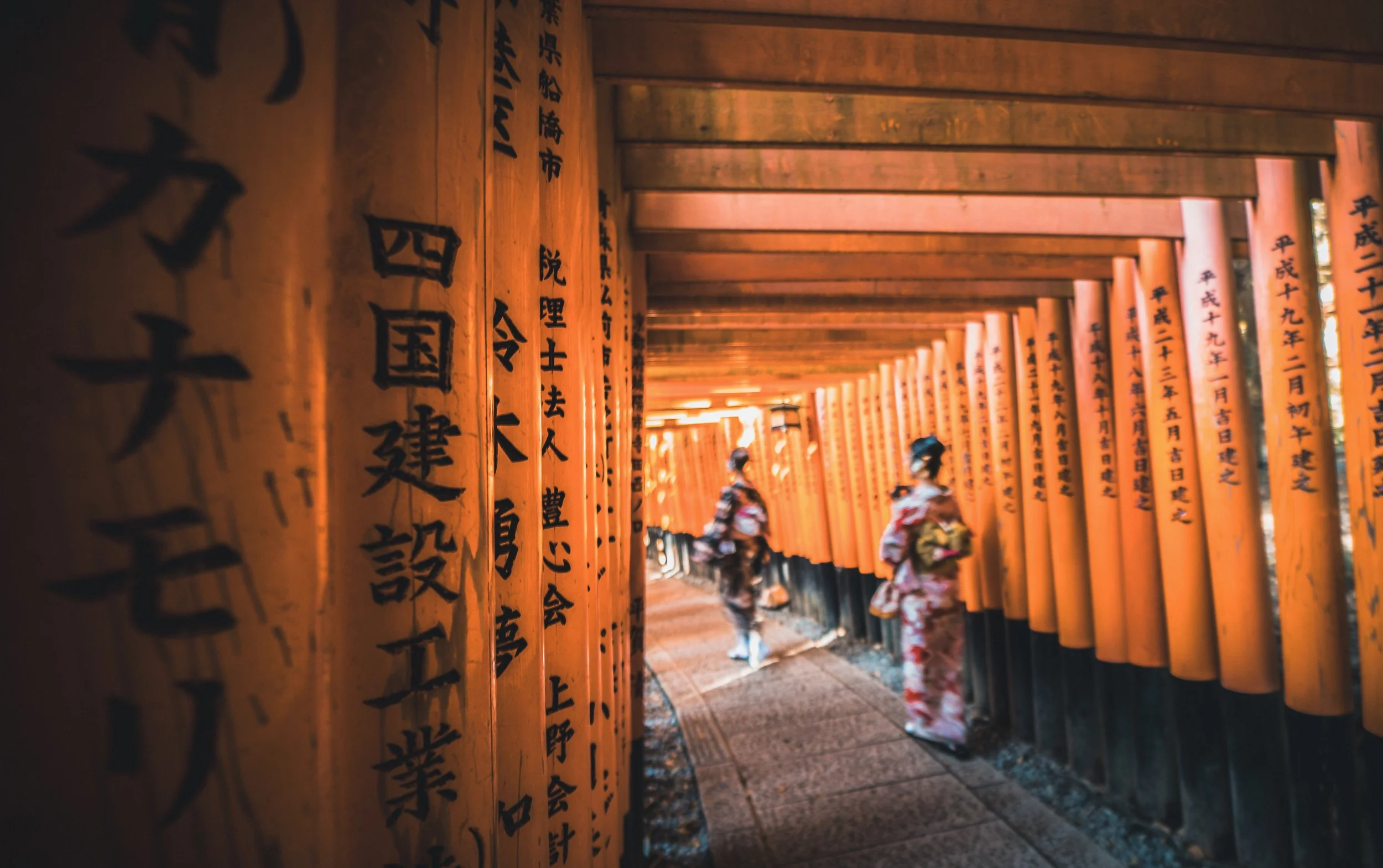 Two women in traditional Japanese kimonos walking through a pathway lined with orange torii gates with black Japanese inscriptions.