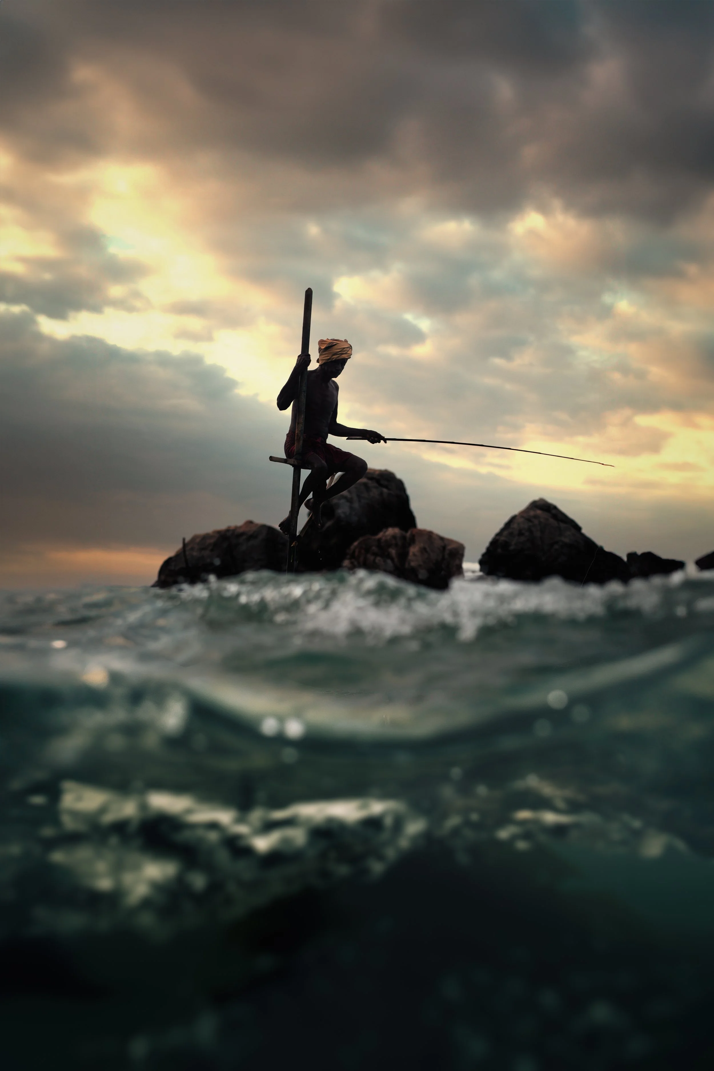 Silhouette of a man fishing on rocks in the ocean during sunset, with a cloudy sky overhead.