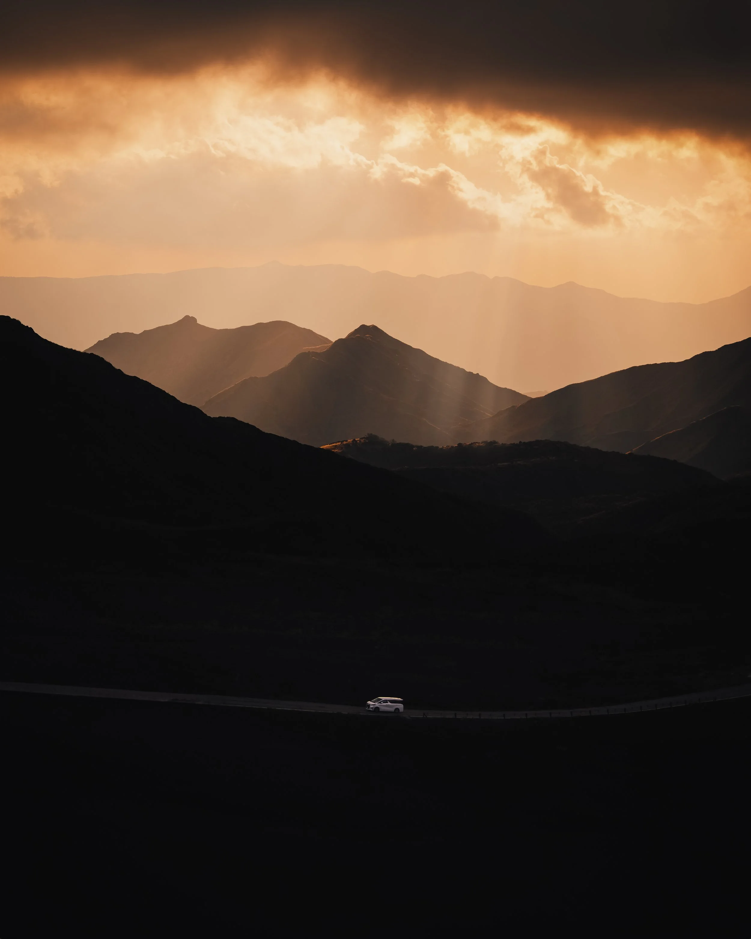 A white car on a winding road through dark mountain valleys under a cloudy sky with sunlight breaking through, illuminating the distant mountain ranges.