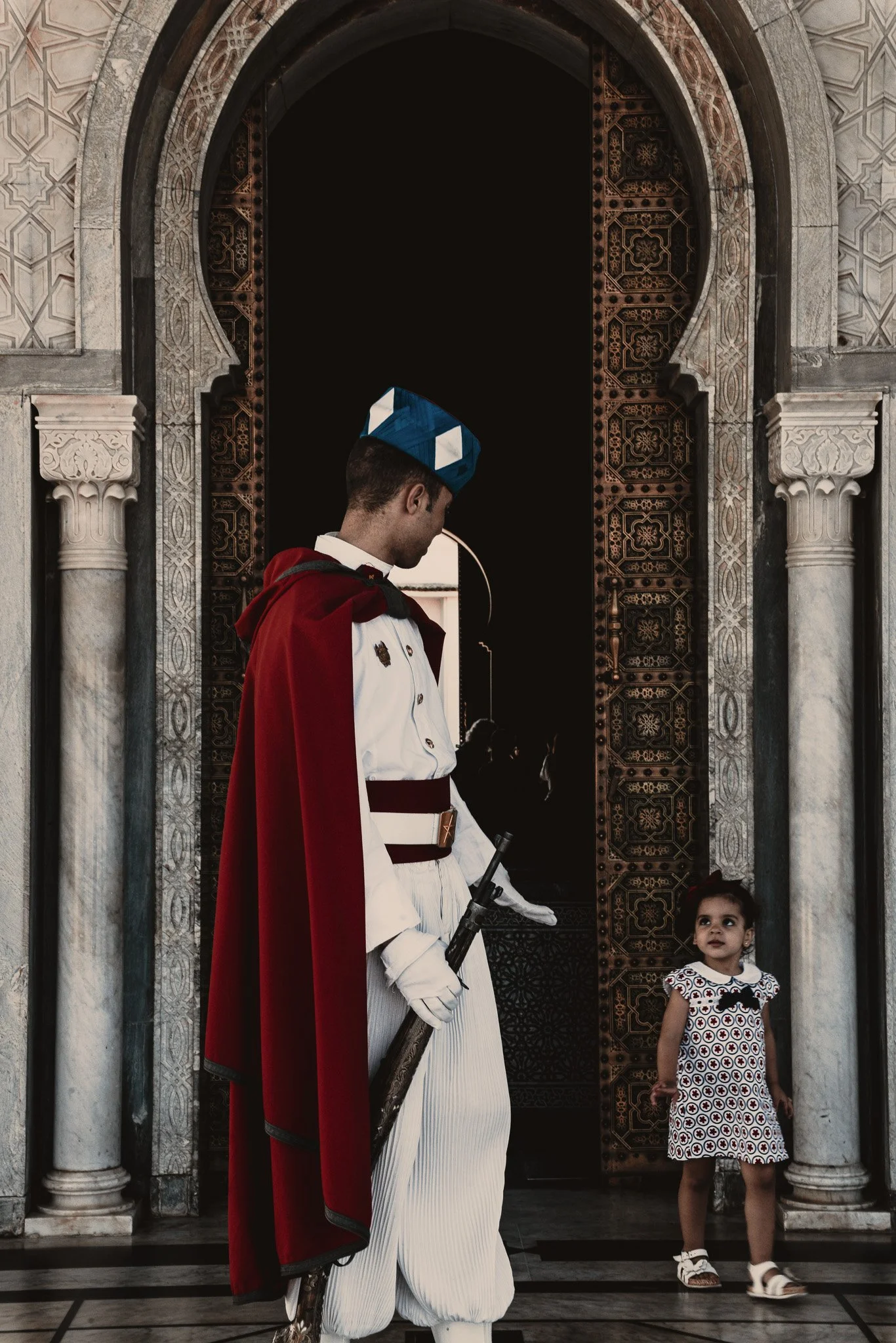 A soldier in a white uniform with a red cape and a blue and white hat stands holding a rifle, looking down at a young girl in a patterned dress, inside a grand building with marble pillars and intricate arches.