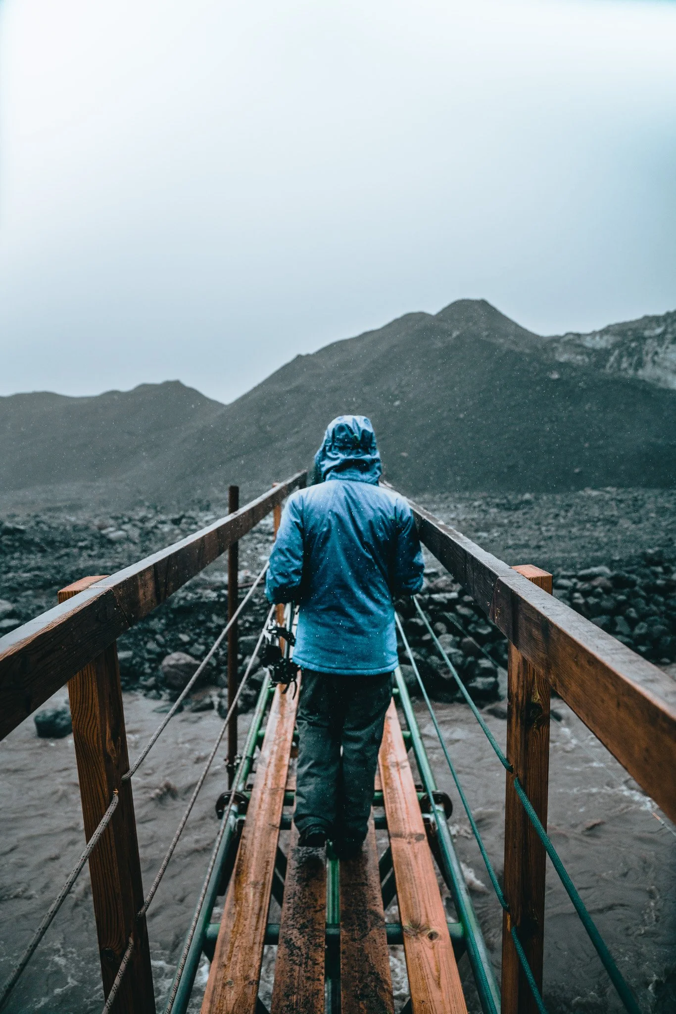 Person wearing a blue raincoat walking on a wooden bridge over rushing water in a mountainous landscape on a rainy day.