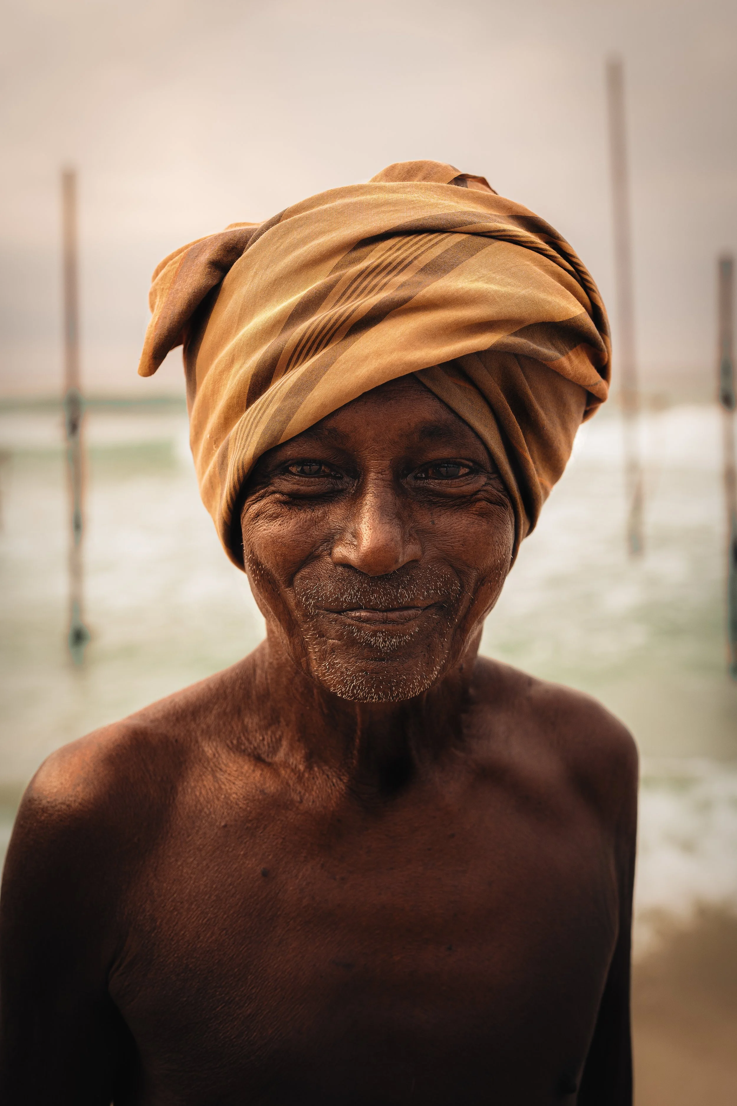 An elderly man with dark skin and a weathered face, wearing a brown and tan headscarf, standing shirtless in front of a body of water.