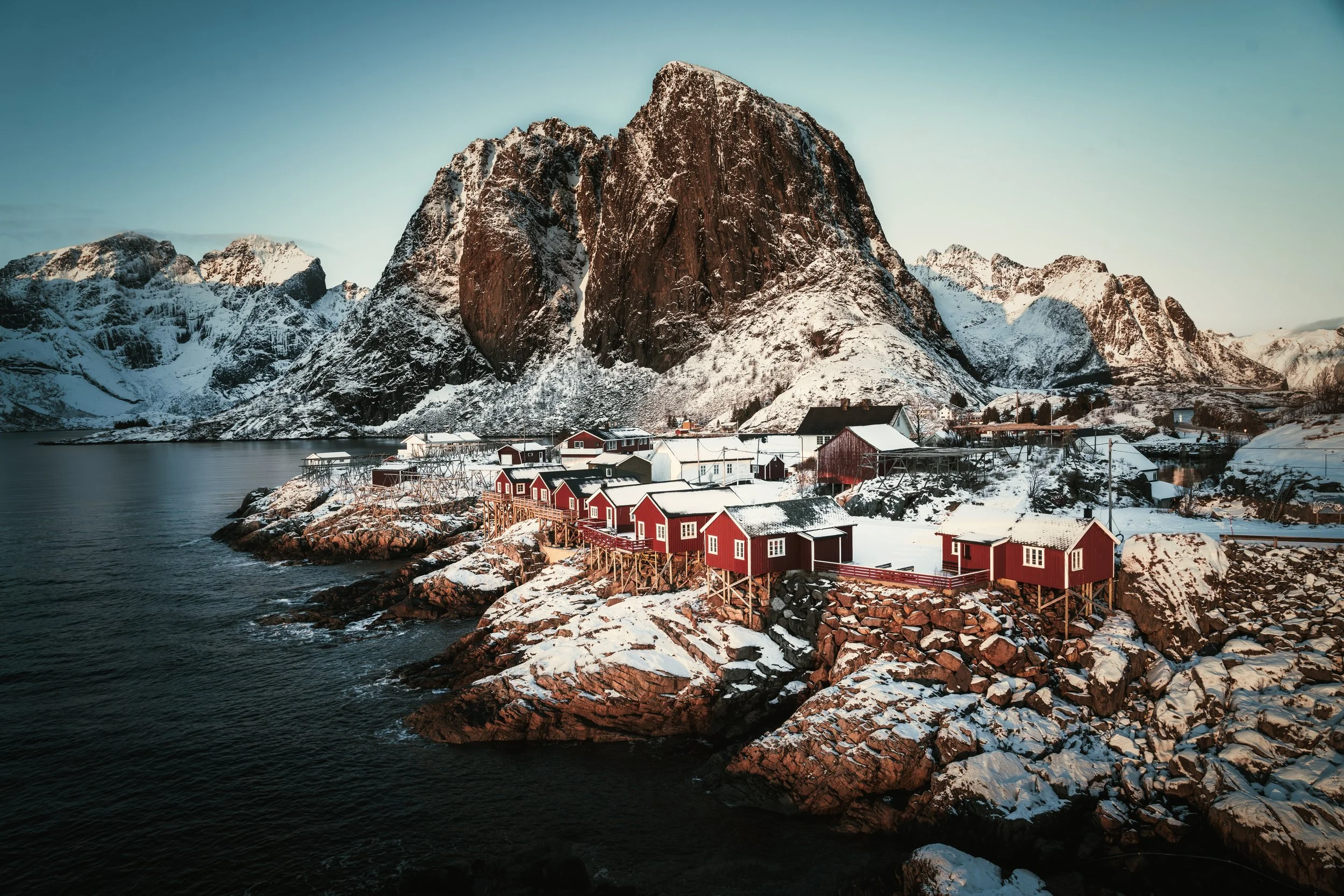 Red cabins beside the water below snowy mountains in Hamnøy, Lofoten, Norway.