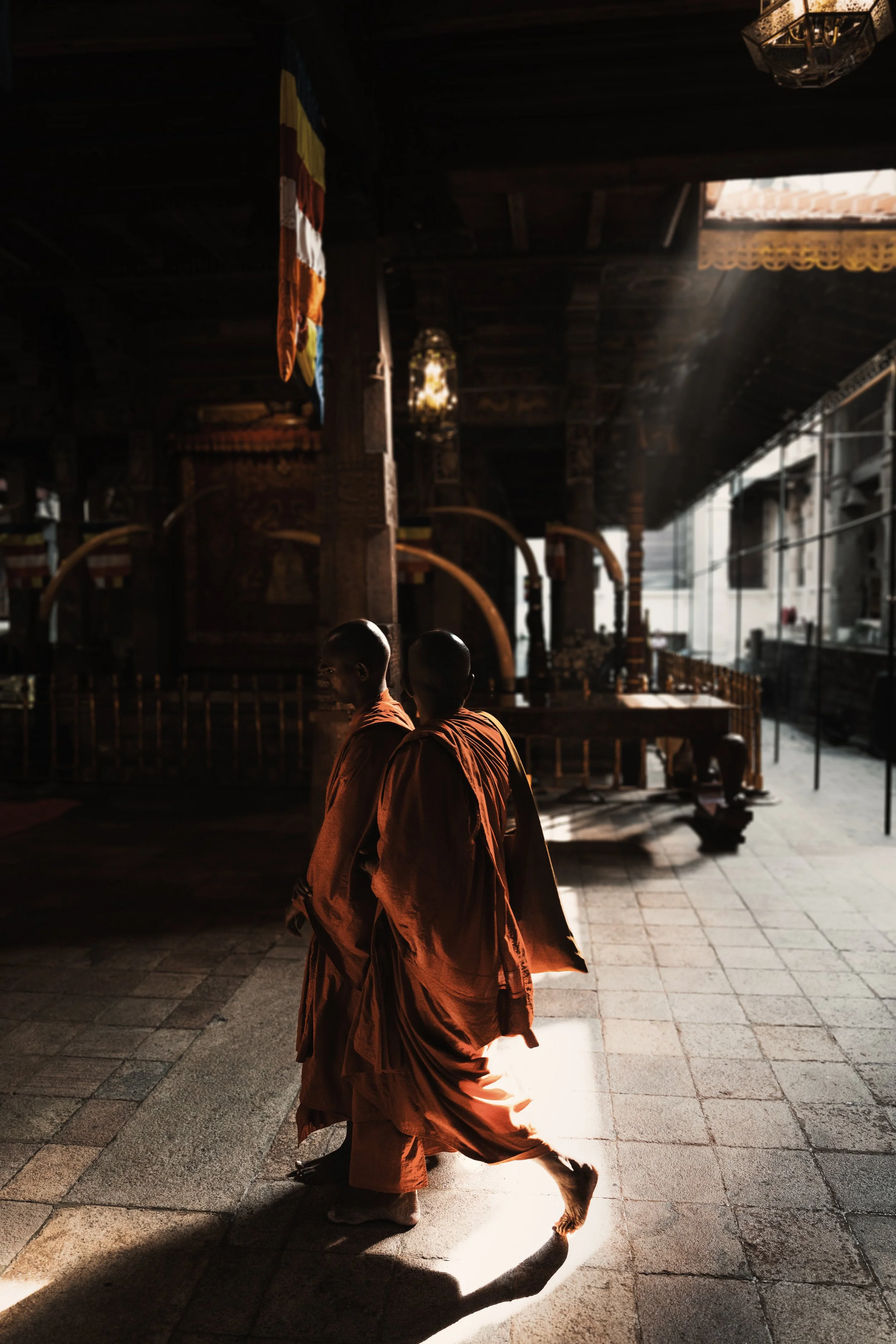 Two monks walking inside a dimly lit temple with sunlight streaming through an open window, traditional decorations hanging, wooden beams, and a prayer area in the background.