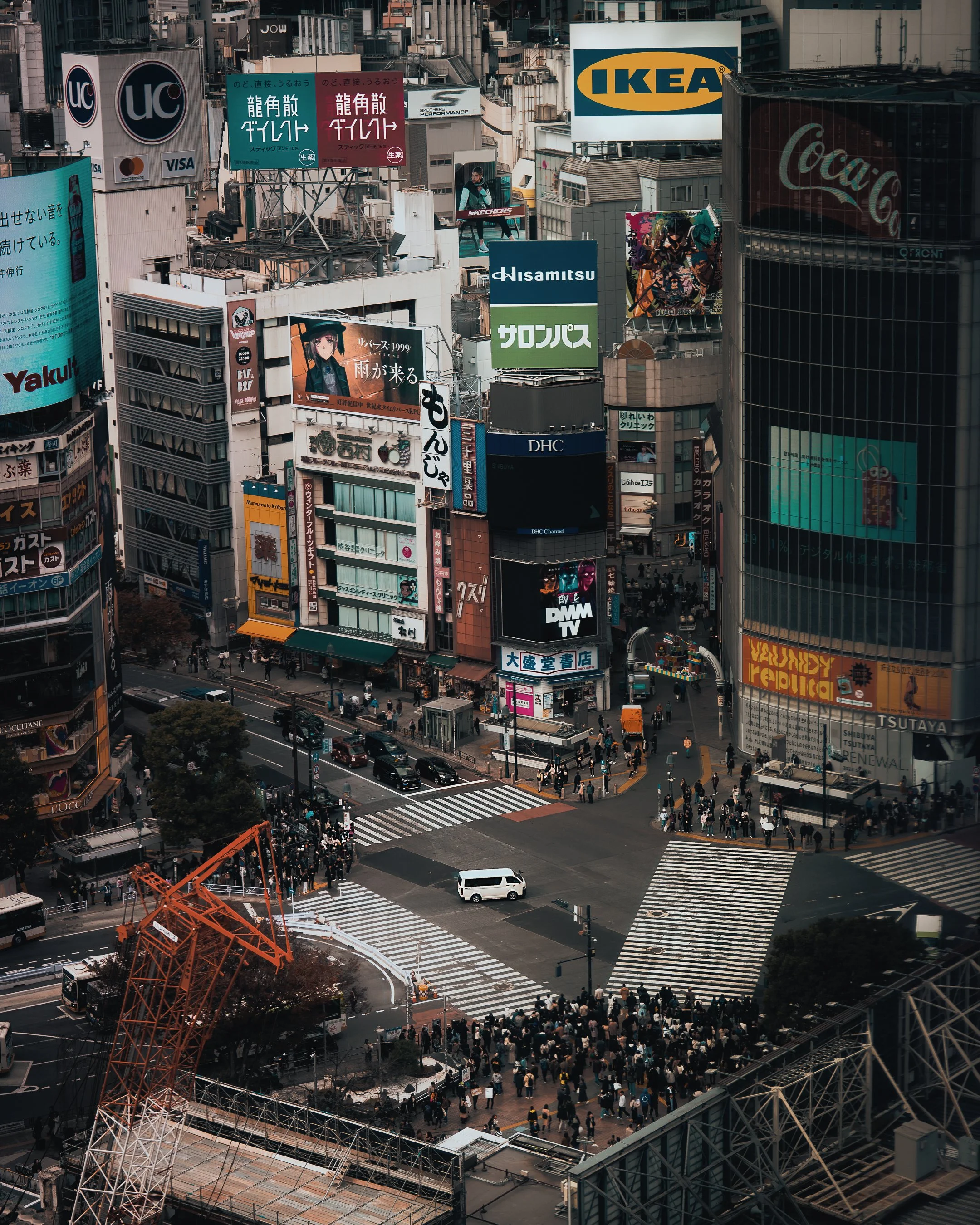 An aerial view of a busy city intersection in Tokyo with pedestrians, cars, and numerous billboards and advertisements, including IKEA, Coca-Cola, and Sony.