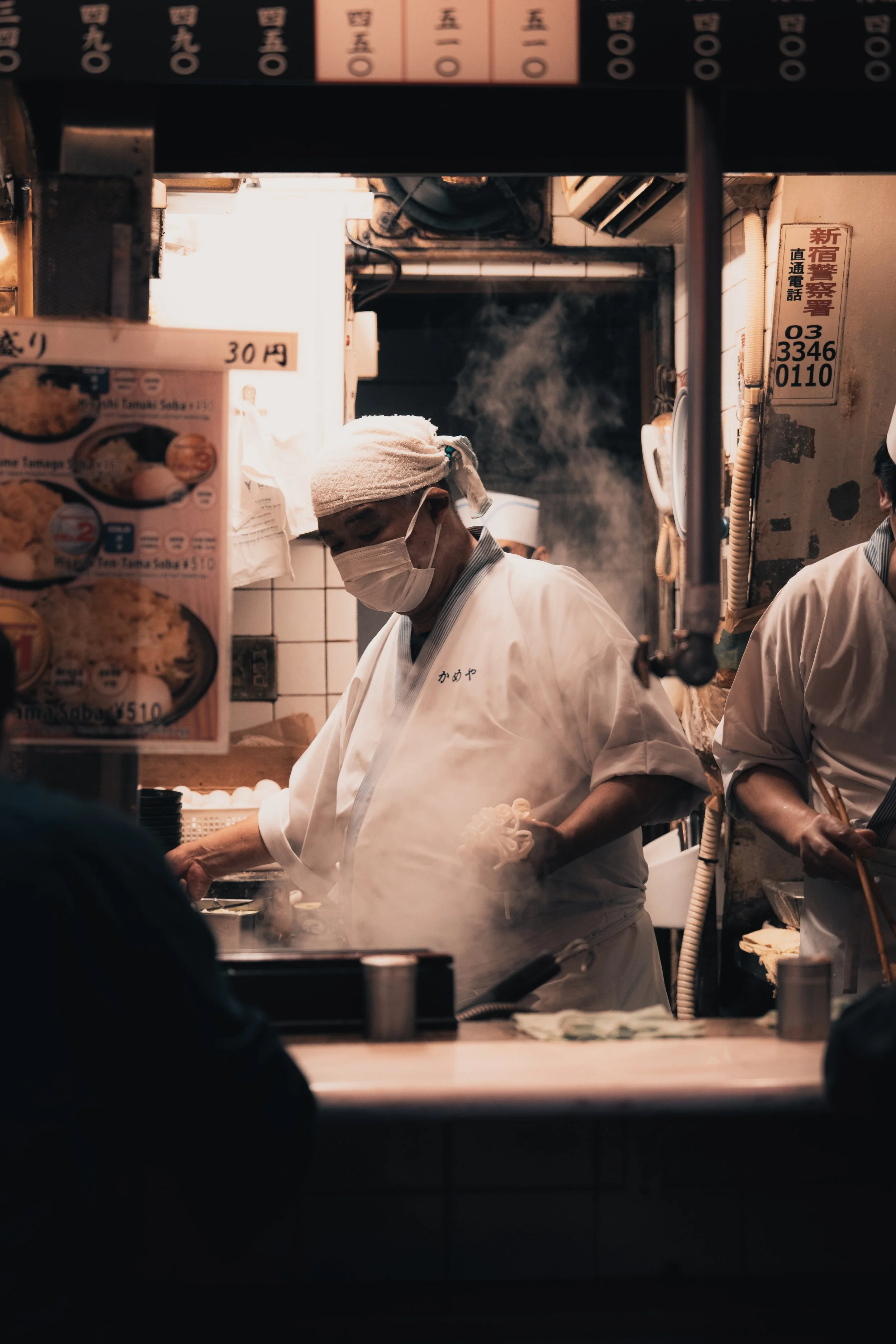 A Japanese chef wearing a mask and head covering preparing food in a small restaurant with steam rising around him.