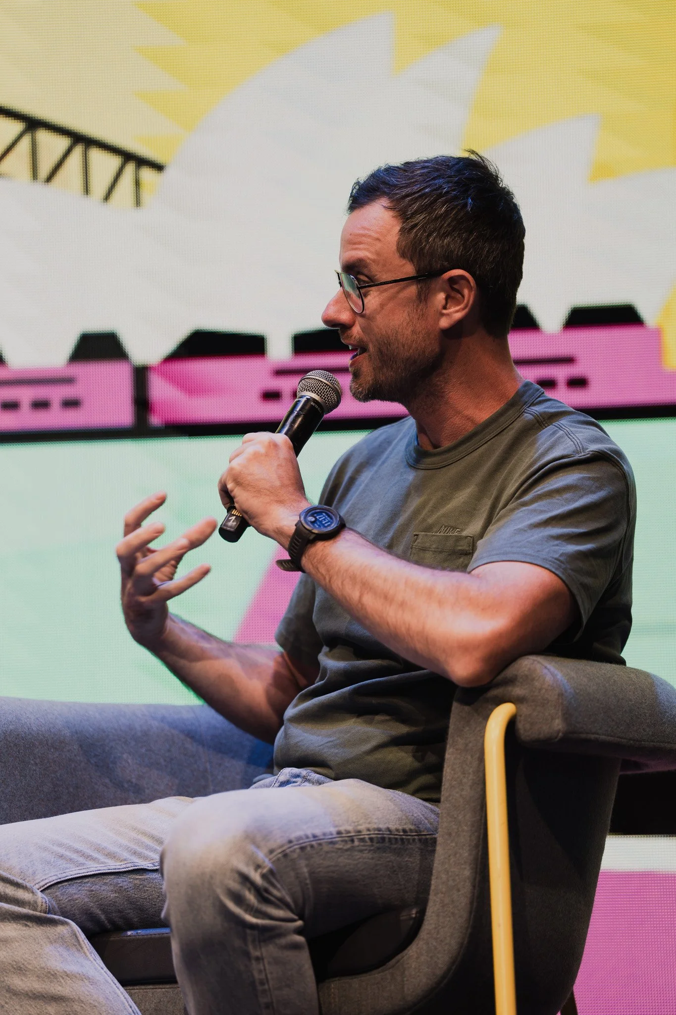 Man with glasses and a beard speaking into a microphone while sitting in a gray chair on a colorful stage.