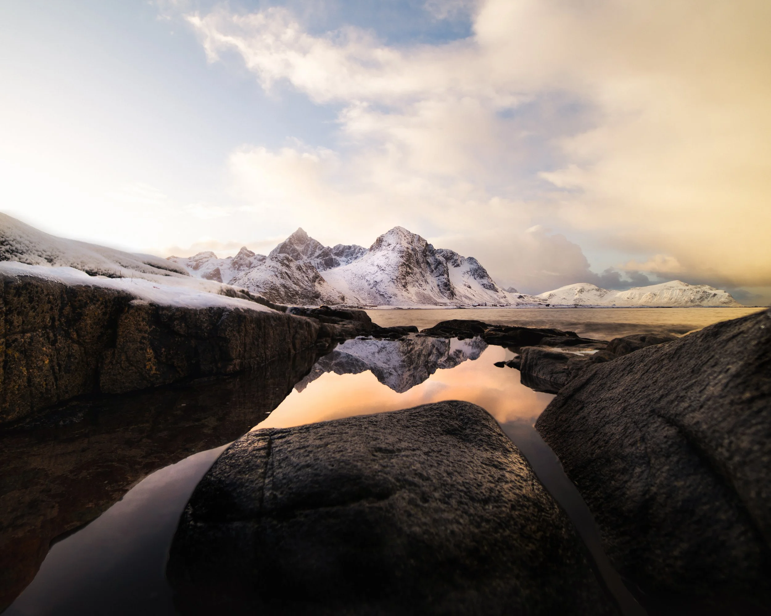 Snow-covered mountains and rocky shoreline reflected in calm water under a partly cloudy sky at sunset.