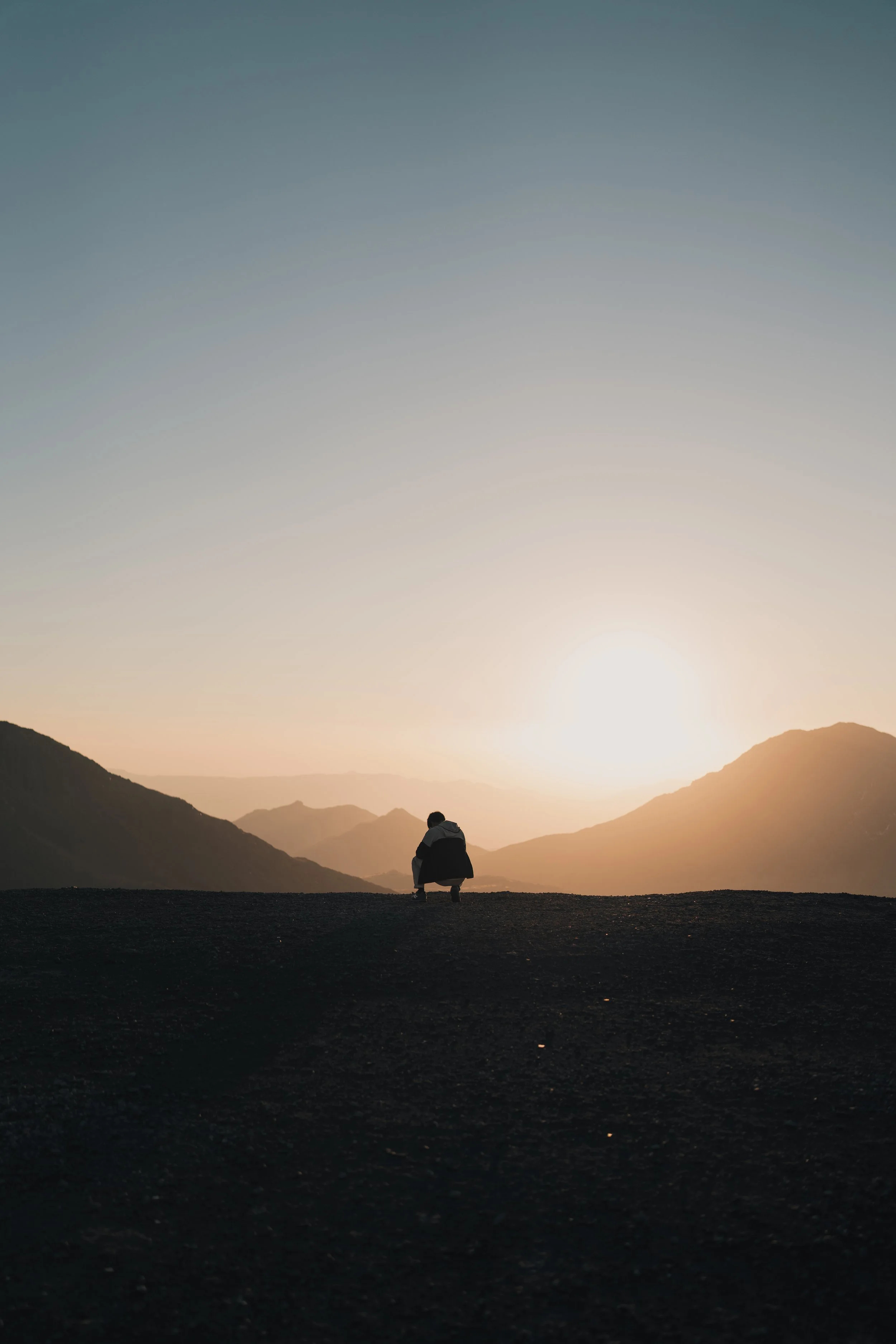 Person sitting alone on a hill at sunset, facing distant mountains with a colorful sky.