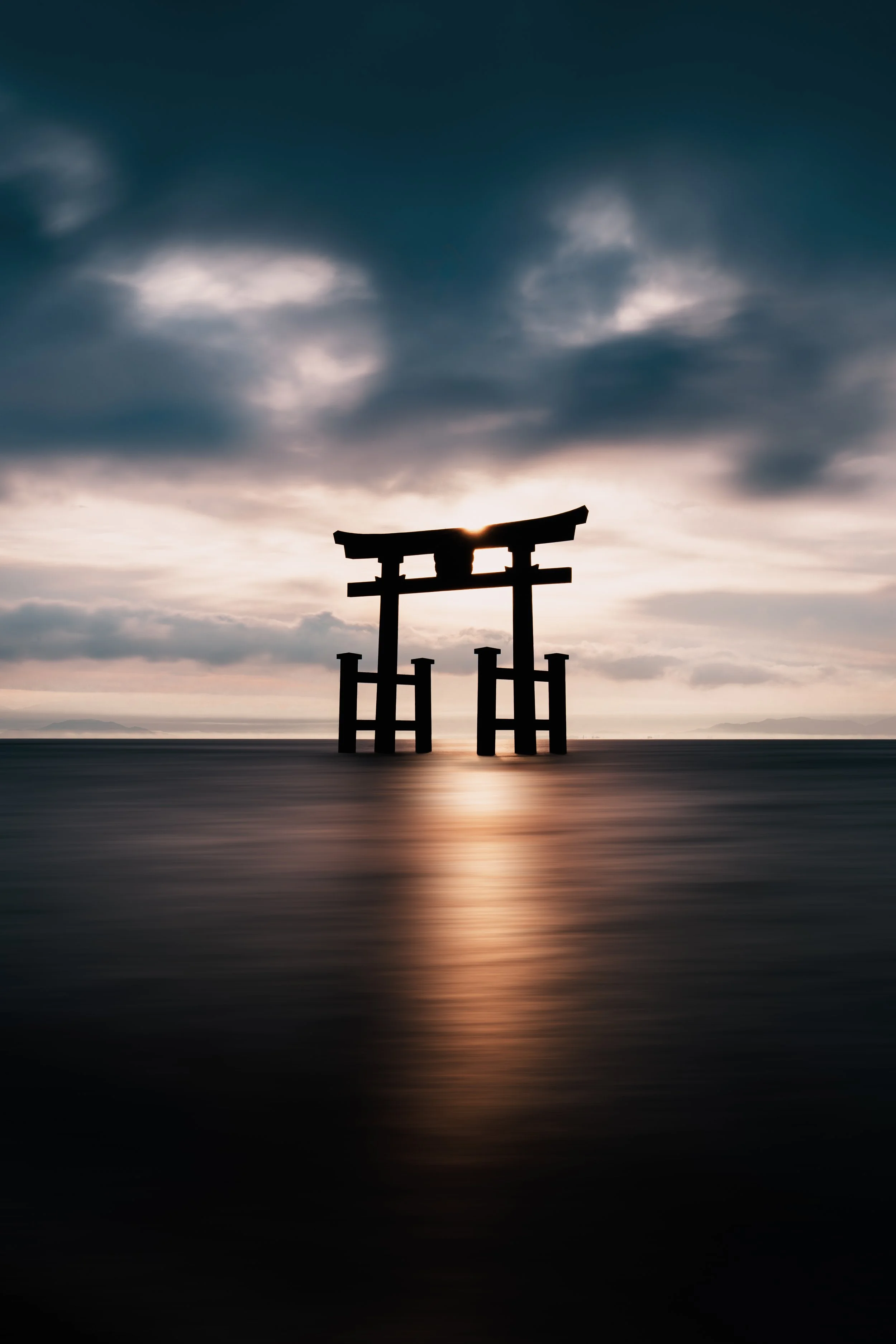 Silhouette of a traditional Japanese torii gate standing in water at sunset or sunrise with dark, cloudy sky.