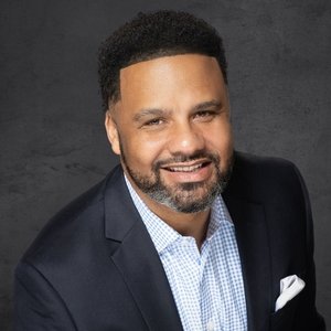 Portrait of a bald man smiling in a dark suit and tie with a black background.
