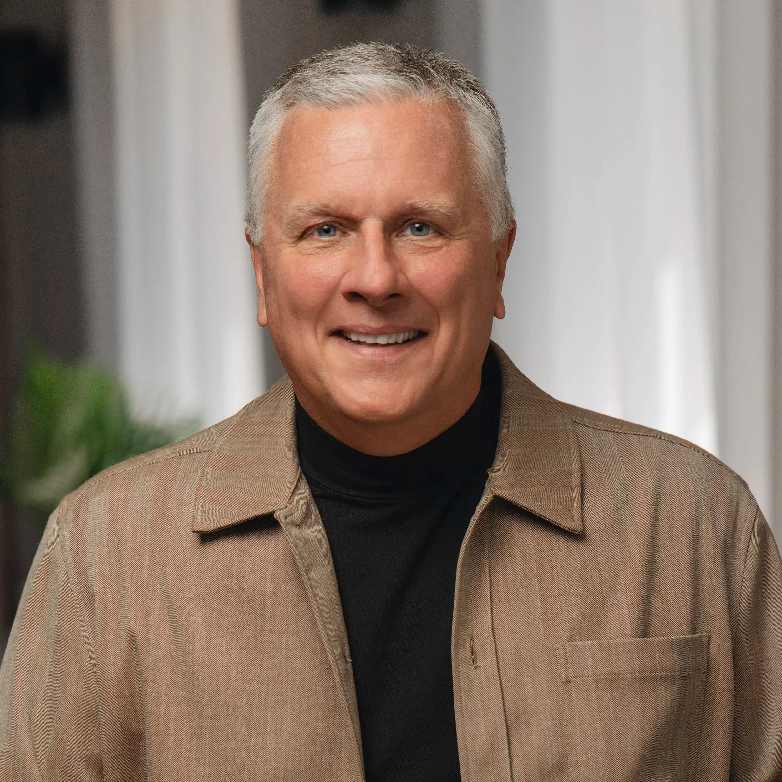 Professional headshot of a Caucasian man white short white hair, in a black shirt and tan jacket