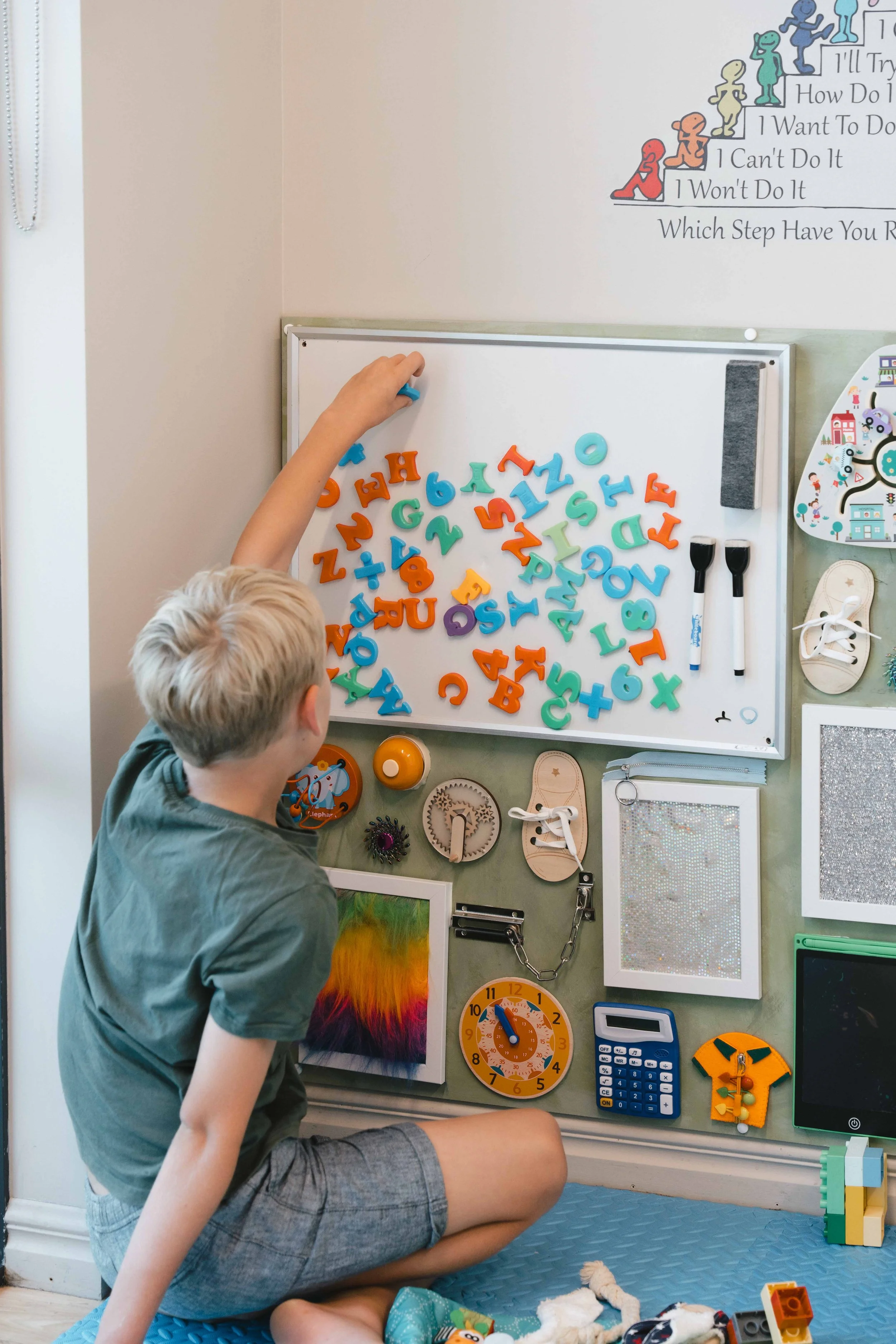 Child playing with colorful alphabet magnets on a whiteboard surrounded by a variety of educational tools and toys, including locks, a clock, and shoes for lacing practice.