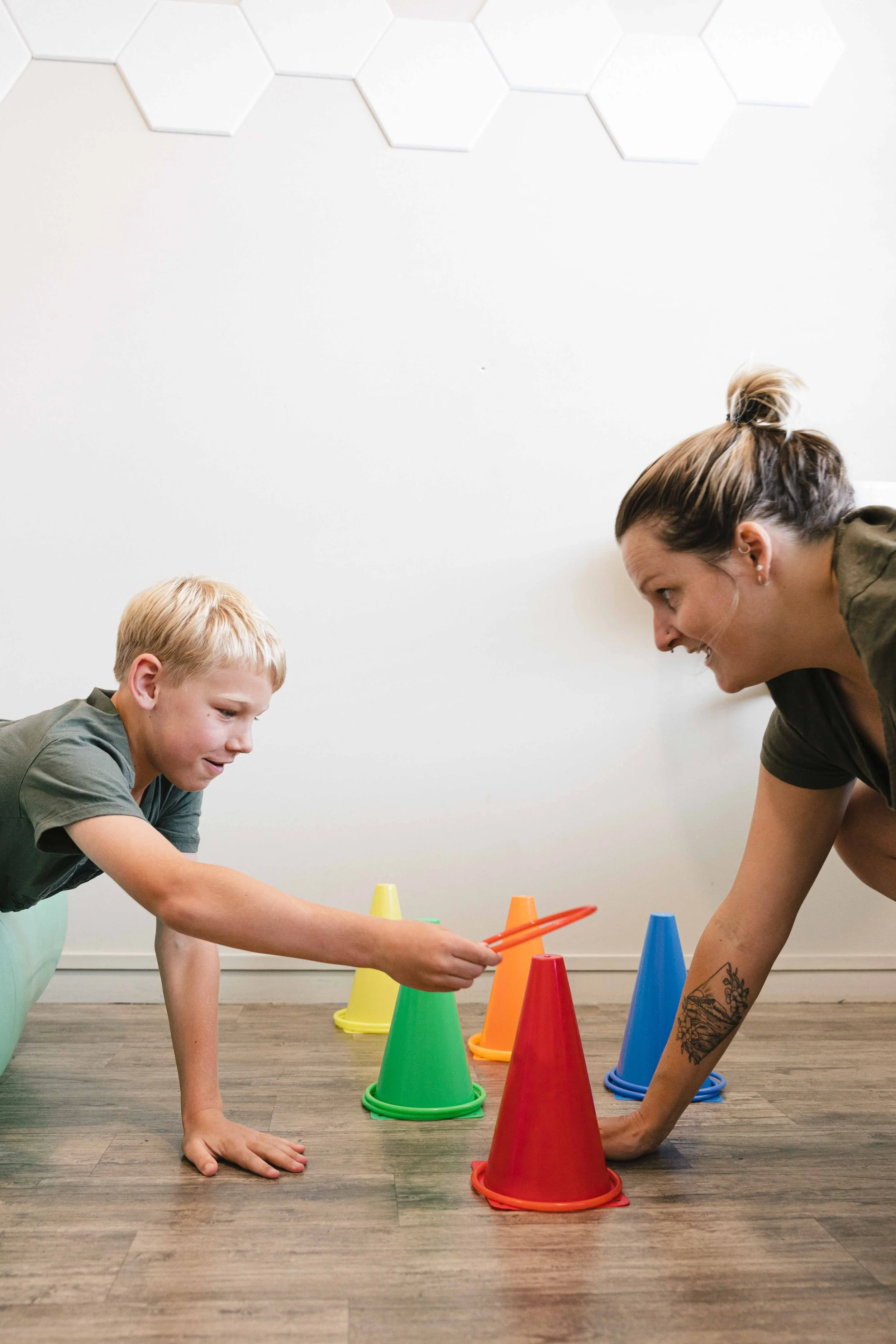 Child and adult playing with colorful cones and ring toss in a room with wooden floor and hexagon wall decor.
