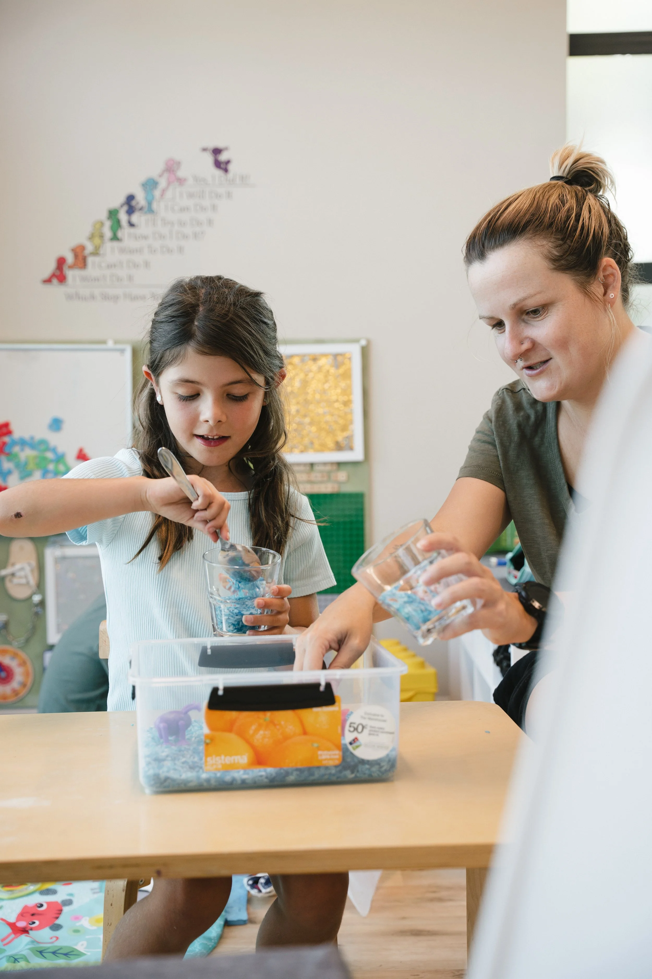 A child and an adult engaging in a classroom activity, using spoons to mix materials in cups, with a storage container on the table. The background features educational wall decor.