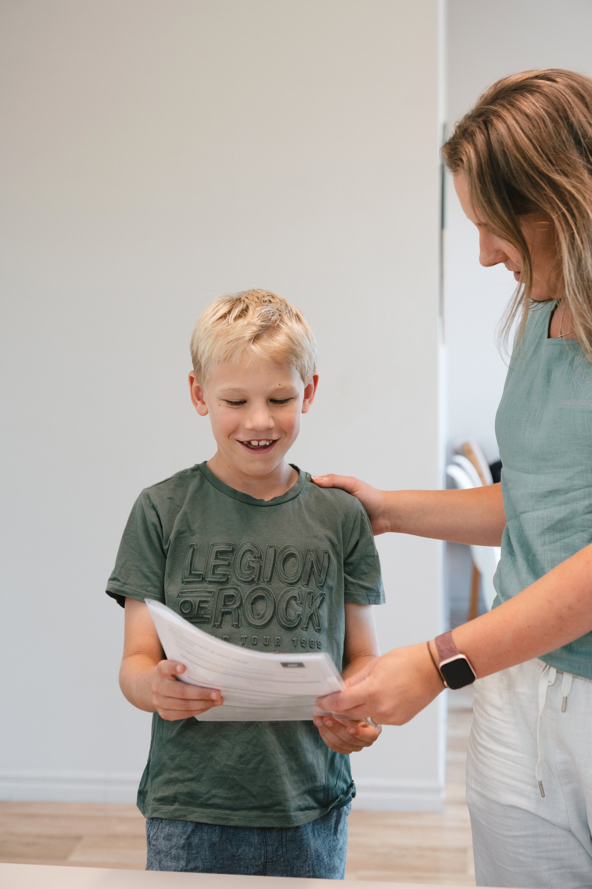 A boy holding a piece of paper and smiling, with a person next to him touching his shoulder, indoors.