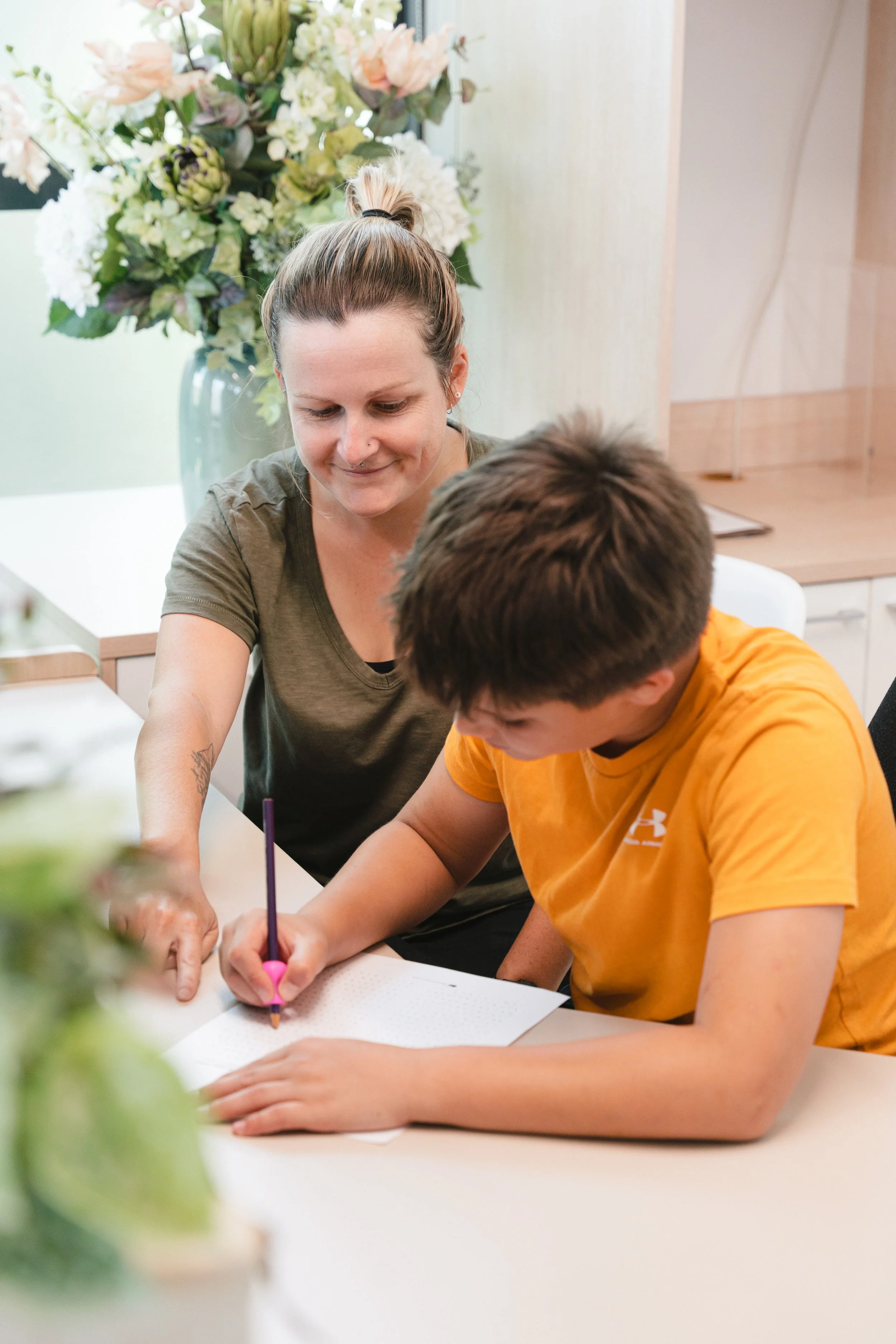Woman helping boy with homework at a table, surrounded by flowers and plants.